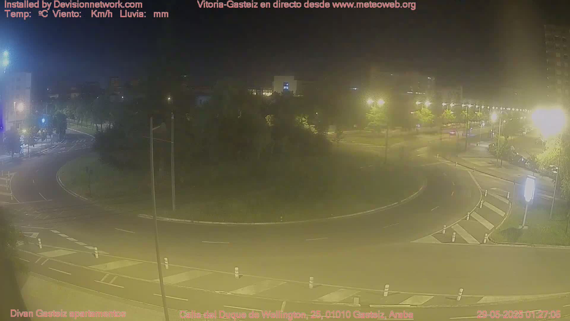 A nighttime view of a traffic roundabout with surrounding buildings and streetlights, under clear skies.