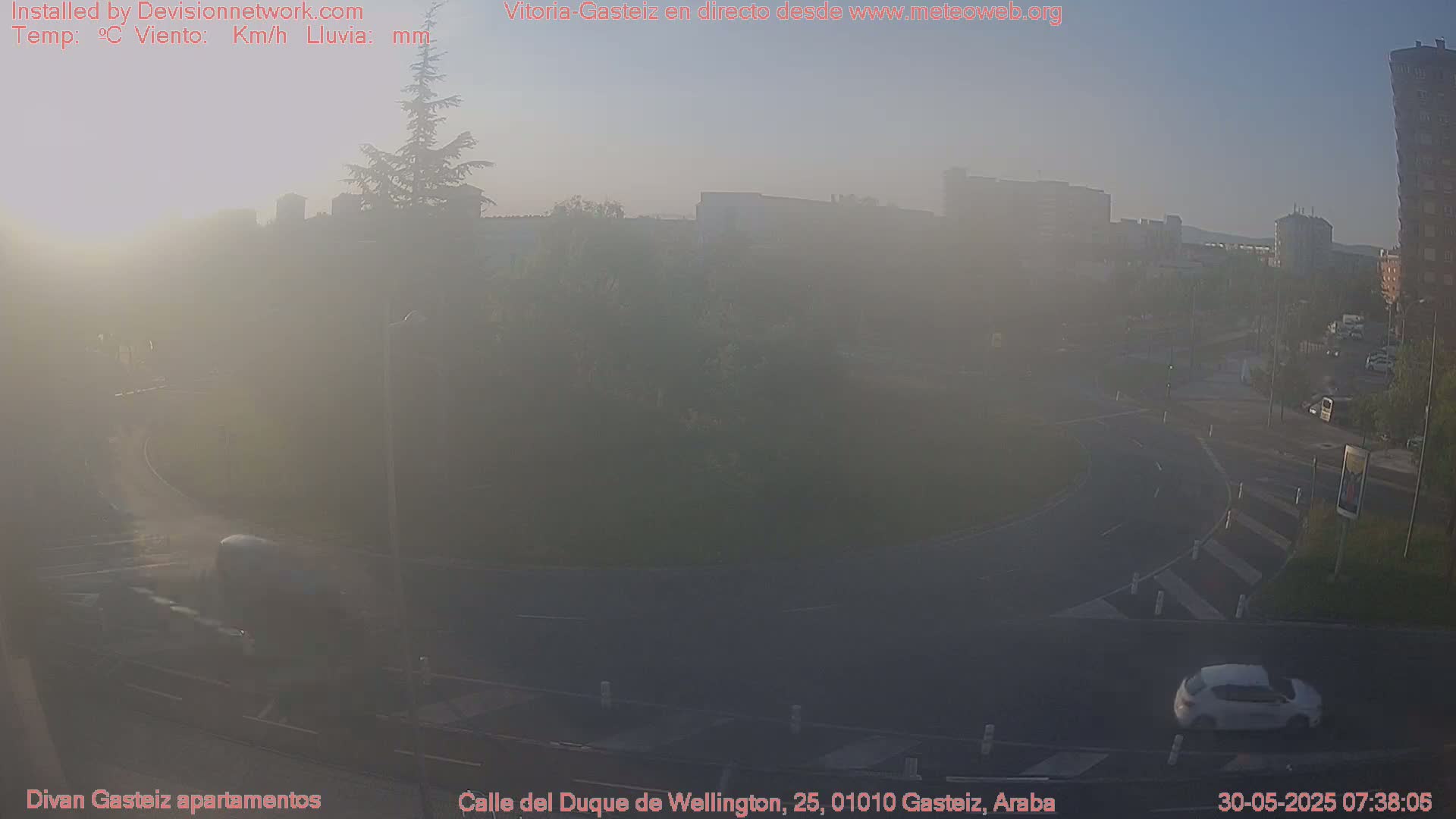 A hazy, early morning scene shows a roundabout and surrounding streets with several cars, some buildings in the distance, and a few trees.