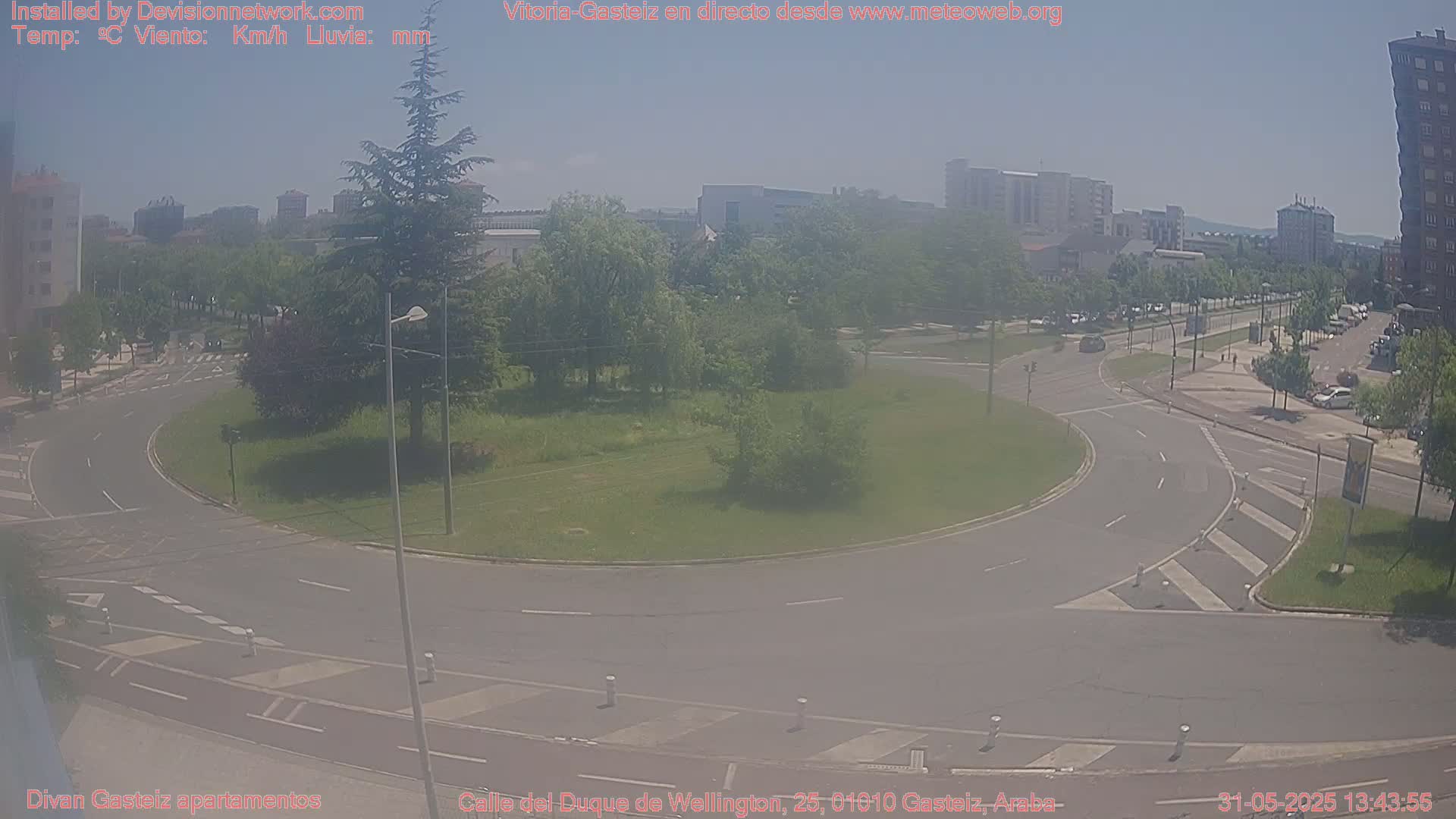 A hazy, sunny day overlooking a roundabout and city streets, with several cars visible and greenery surrounding the roundabout.