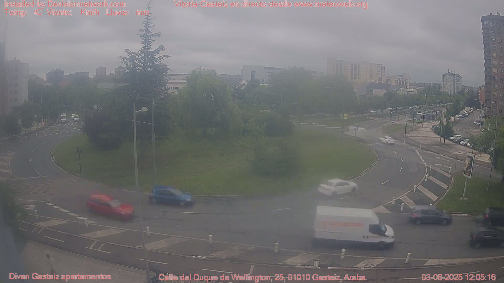 A hazy, overcast day reveals a roundabout with several cars and a van navigating its lanes, surrounded by buildings and greenery.
