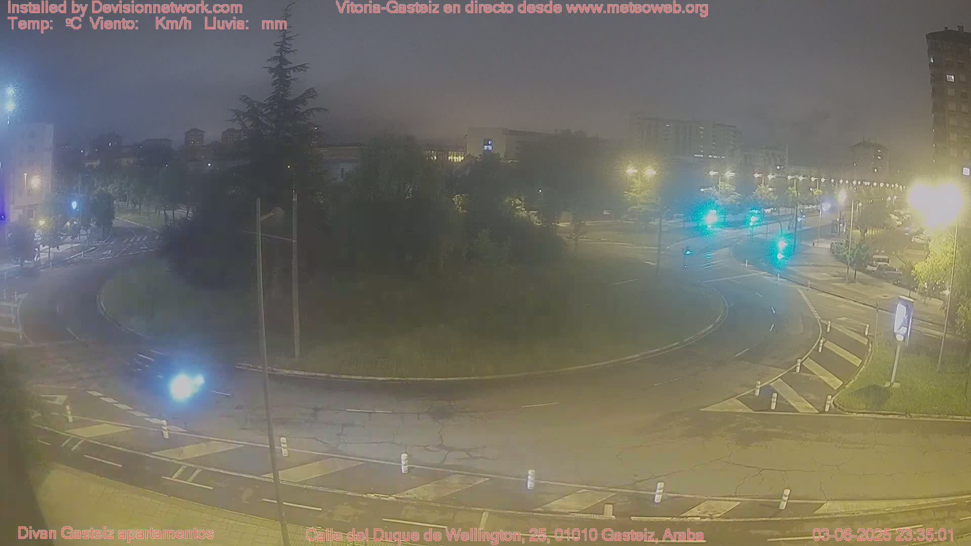 A nighttime, slightly hazy view of a roundabout with several cars, streetlights, and buildings in the background.