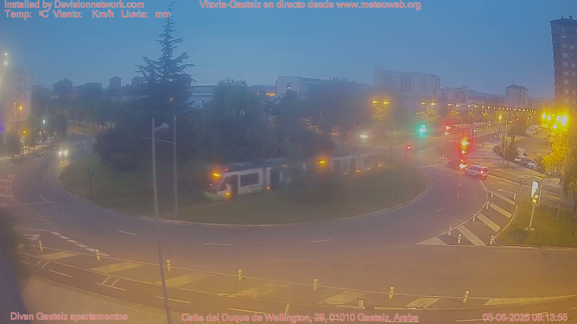 A street scene at dusk shows a tram traversing a roundabout, with cars at the intersection, and buildings and trees in the background.