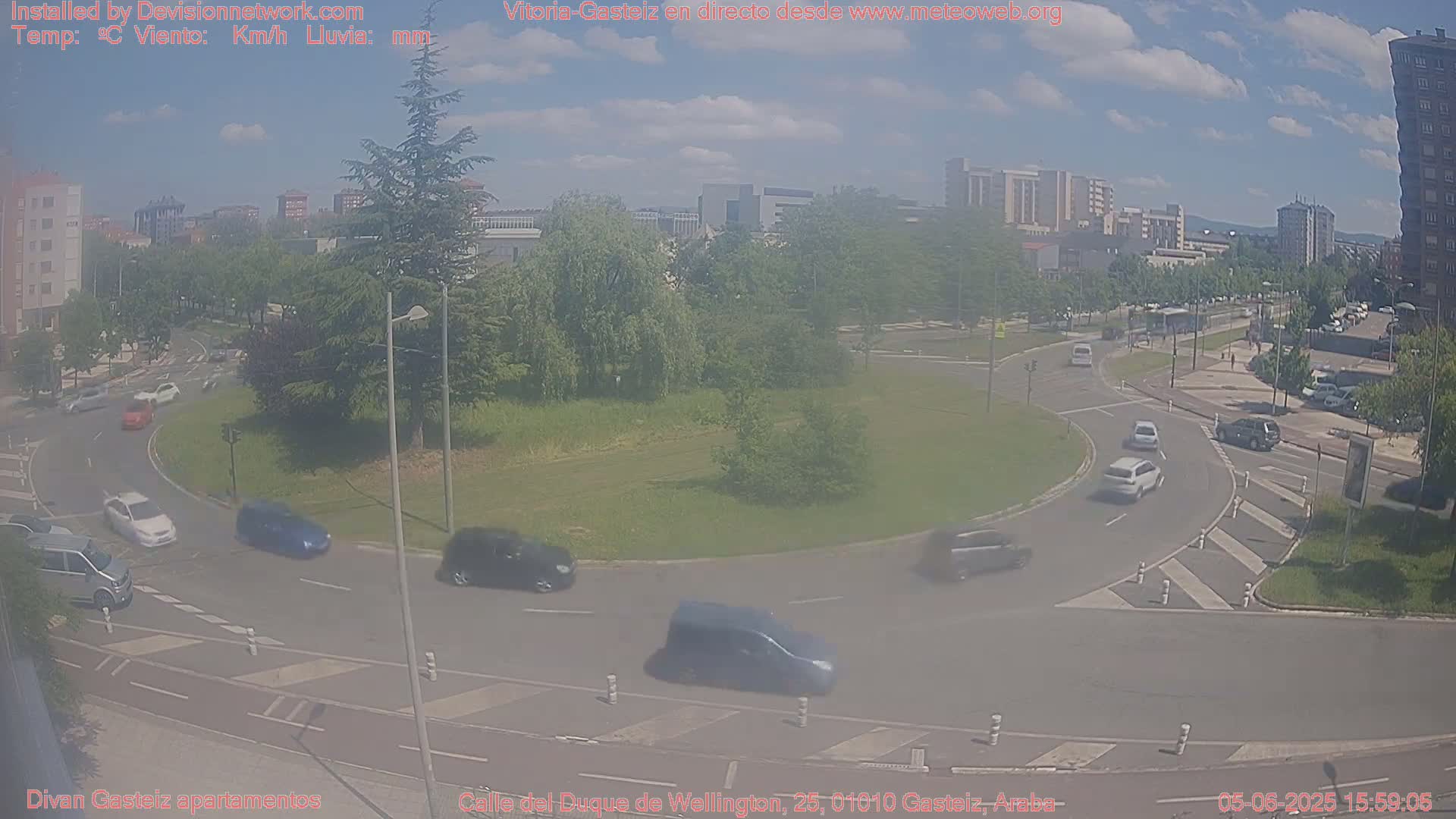A partly cloudy day shows several cars circulating a roundabout in a city, with buildings and trees in the background.