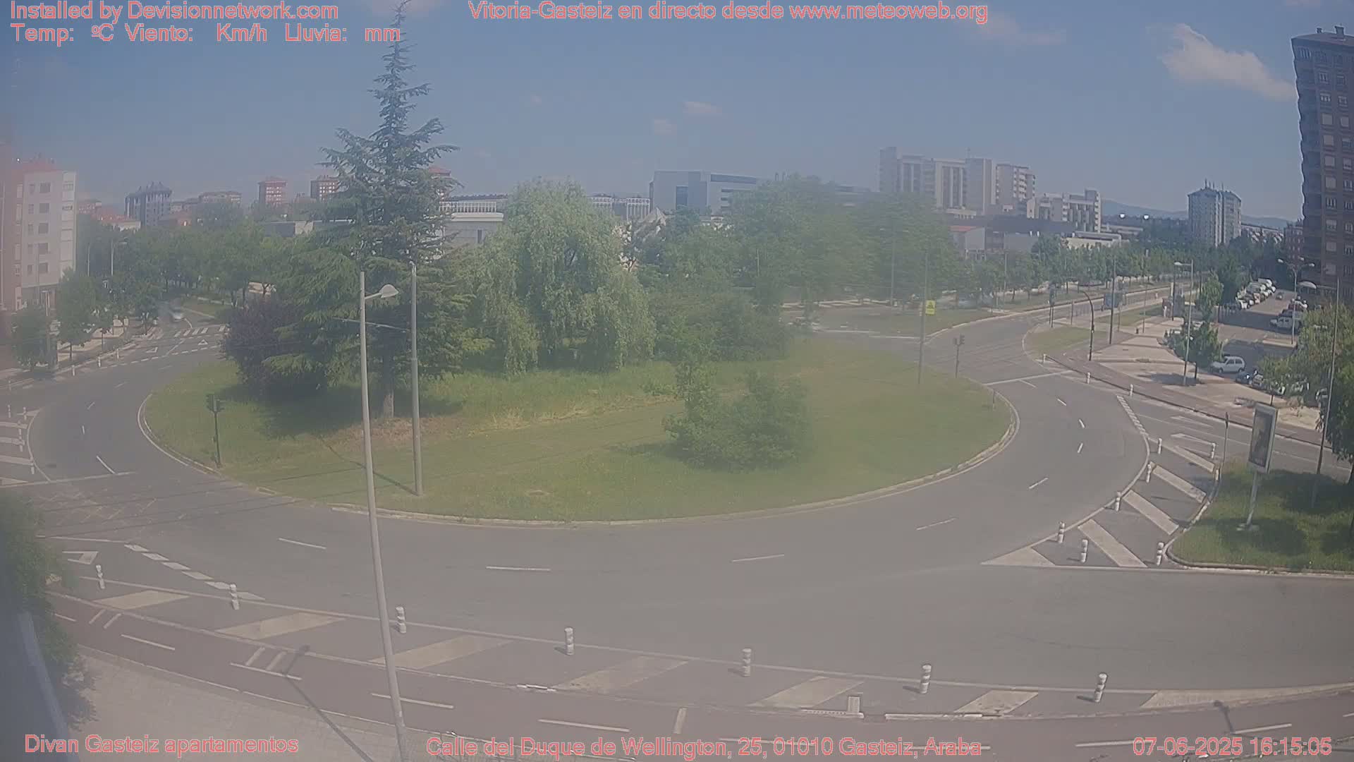 A mostly sunny day shows a roundabout in a city with several buildings in the background and some cars visible on the roads.