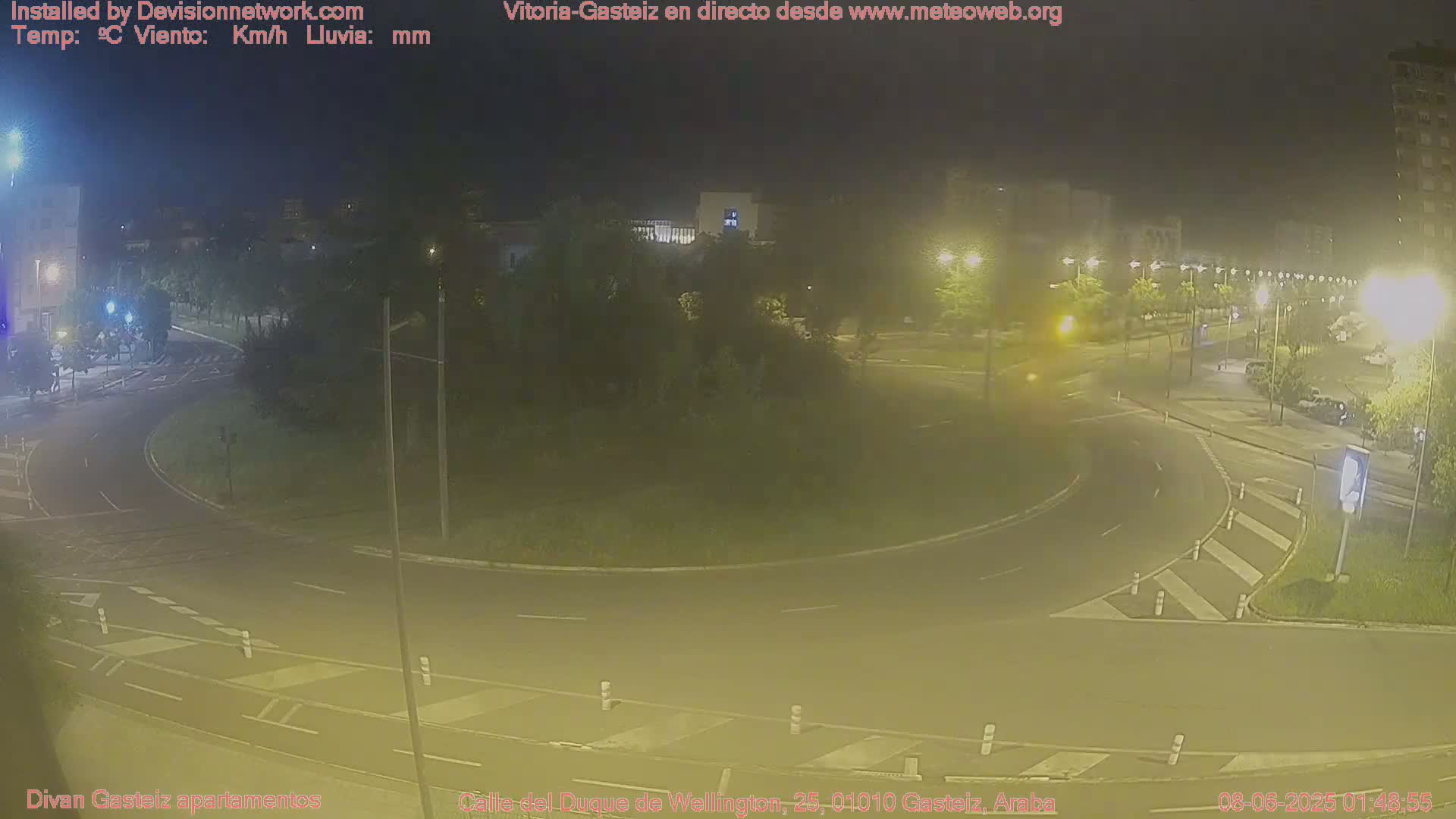 A nighttime, hazy view of a roundabout intersection with surrounding buildings and streetlights.
