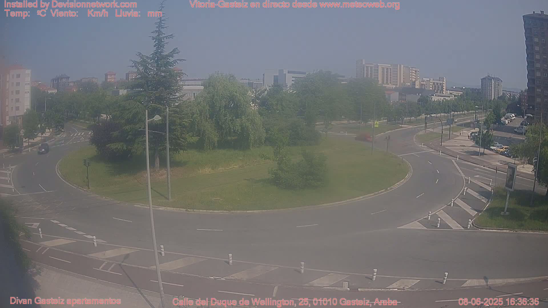 A hazy, sunny day shows a roundabout surrounded by green grass and trees, with a few cars visible on the roads nearby and buildings in the background.