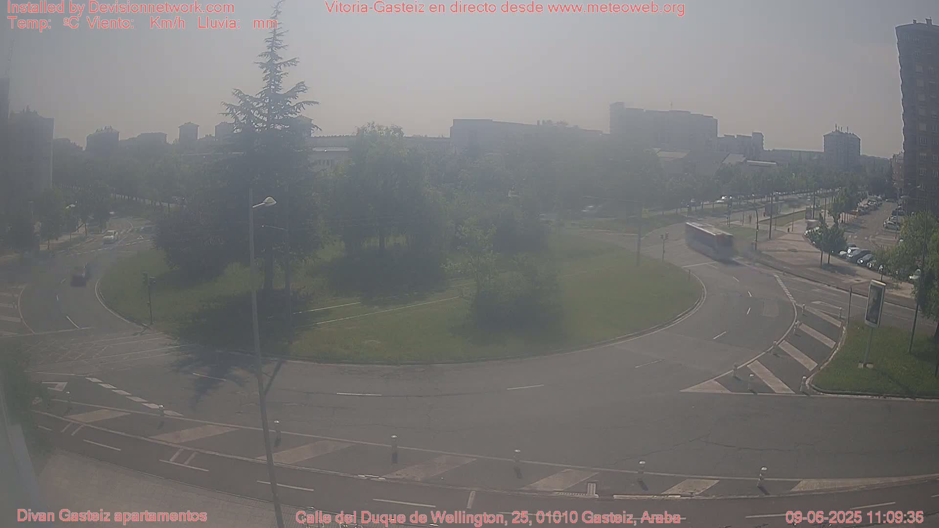 A hazy, sunny day reveals a roundabout with light traffic and a bus, surrounded by greenery and city buildings in the distance.