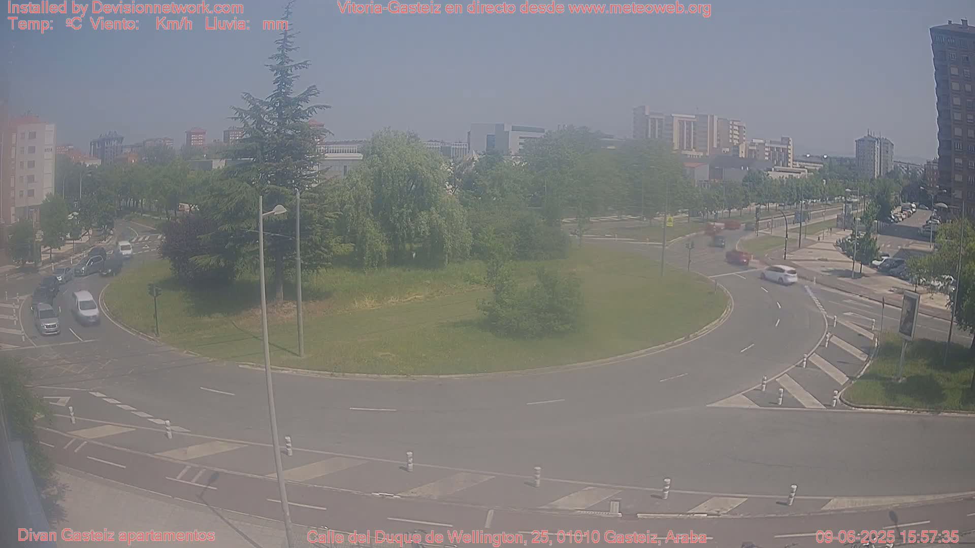 A hazy, sunny day reveals a roundabout with several cars, surrounded by green spaces and buildings in the distance.