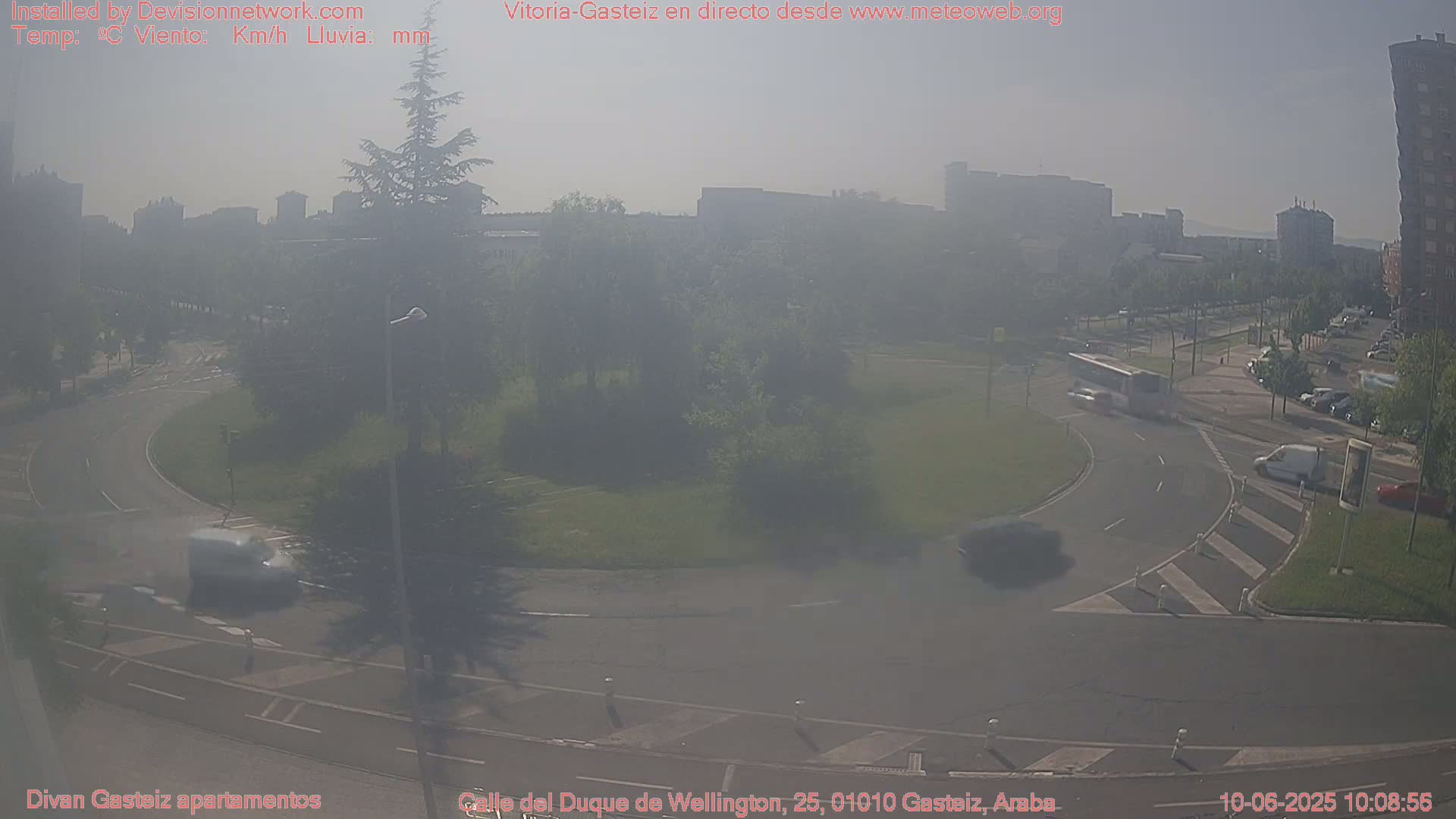 A hazy view of a roundabout with several vehicles, surrounded by trees and buildings in a city.