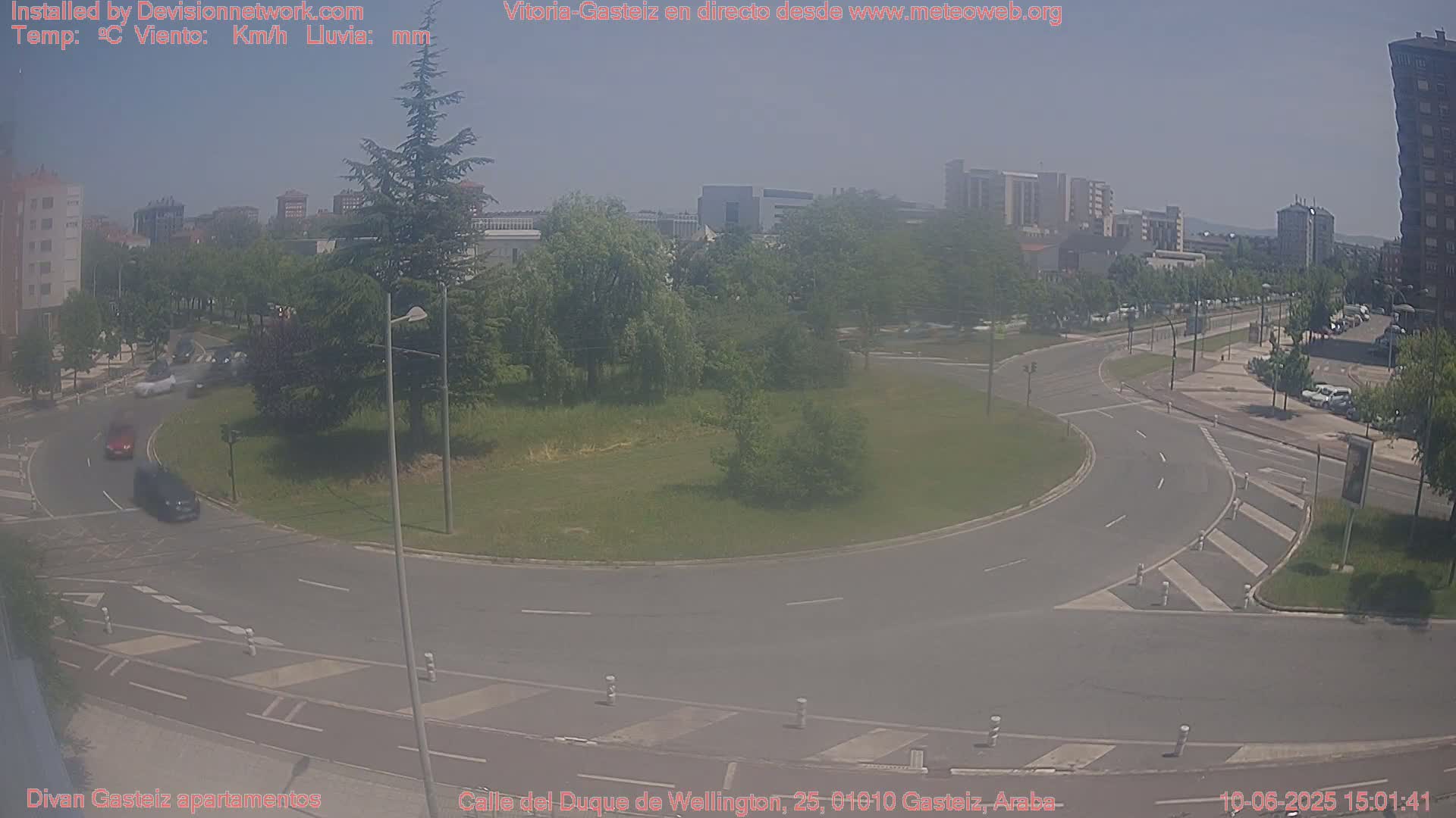 A hazy, sunny day shows a roundabout in a city with several cars driving on the road and buildings visible in the background.