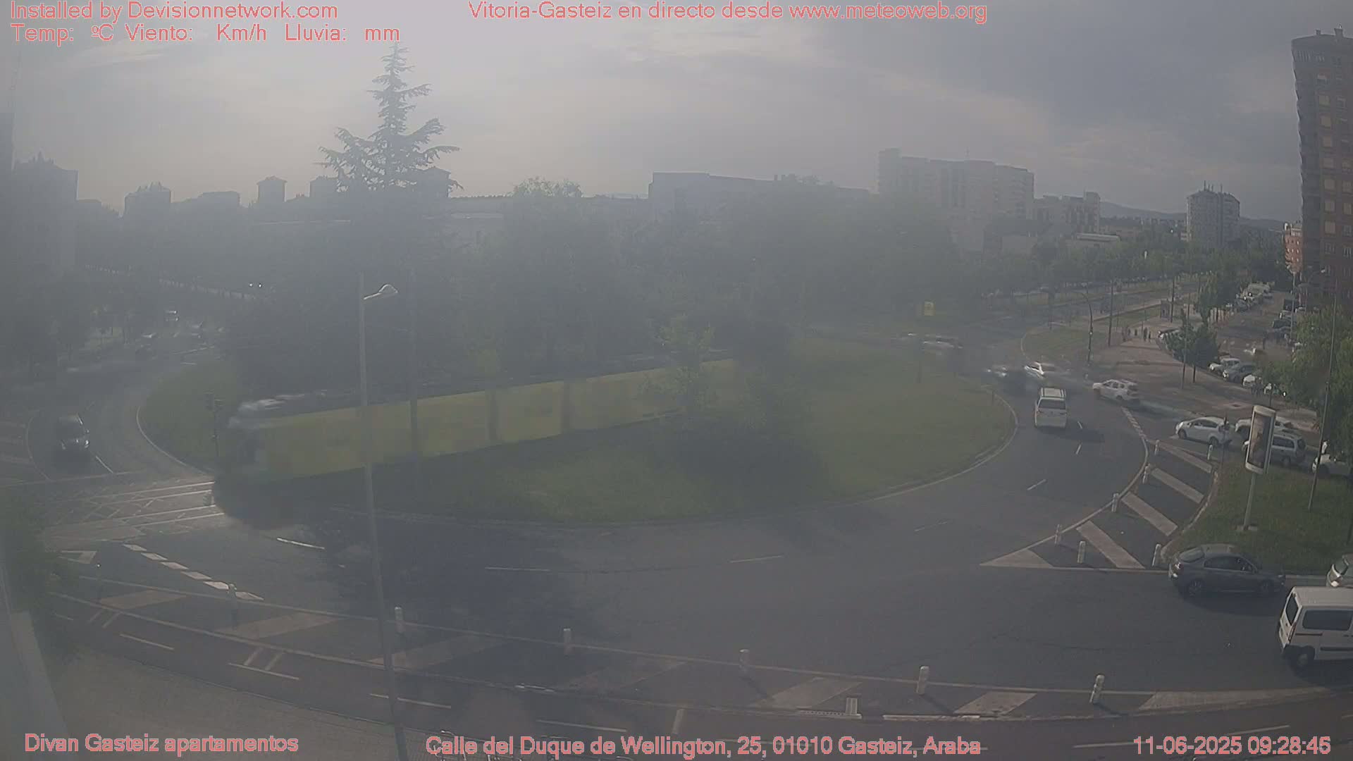 A hazy view of a roundabout in a city, with several cars visible and buildings in the background.