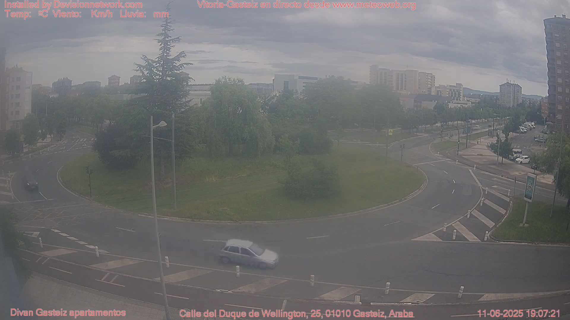 A hazy view of a roundabout with a car driving through it, surrounded by buildings, trees, and overcast skies.