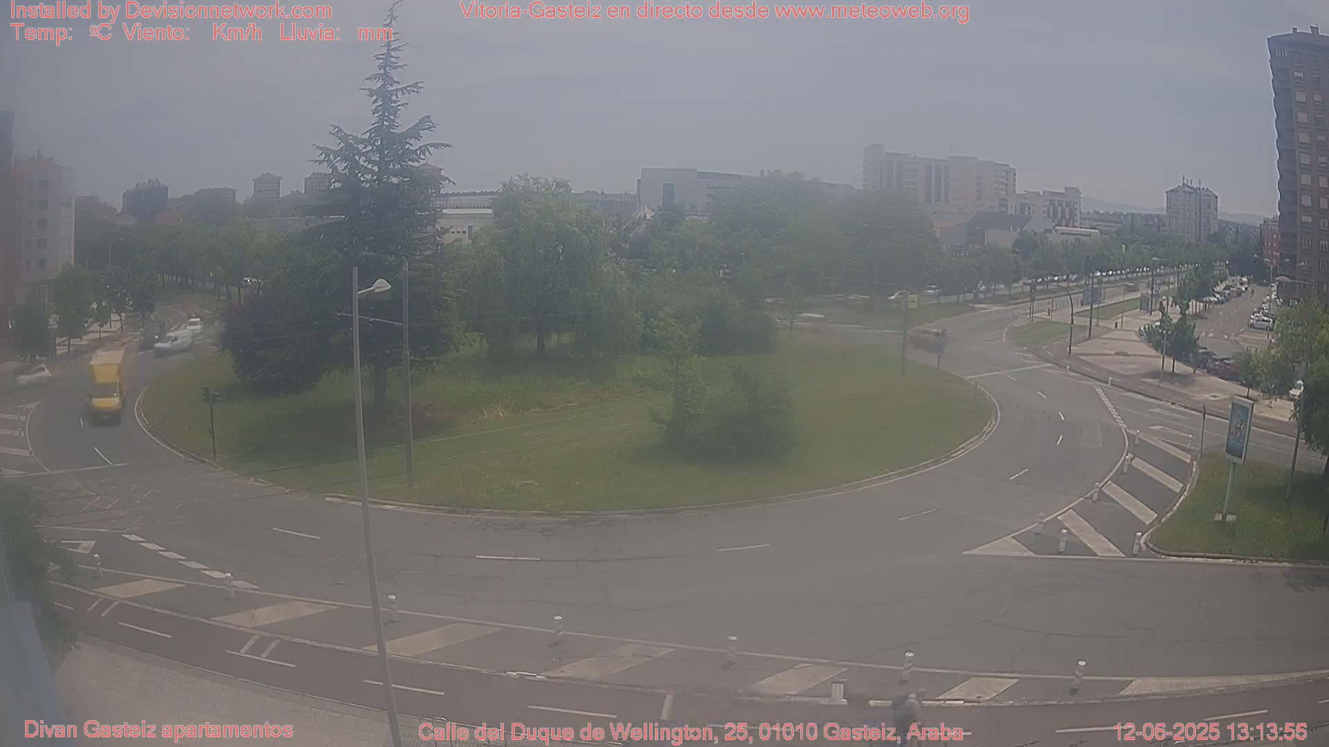 A hazy view of a roundabout and surrounding city streets with several vehicles, trees, and buildings under a mostly clear sky.