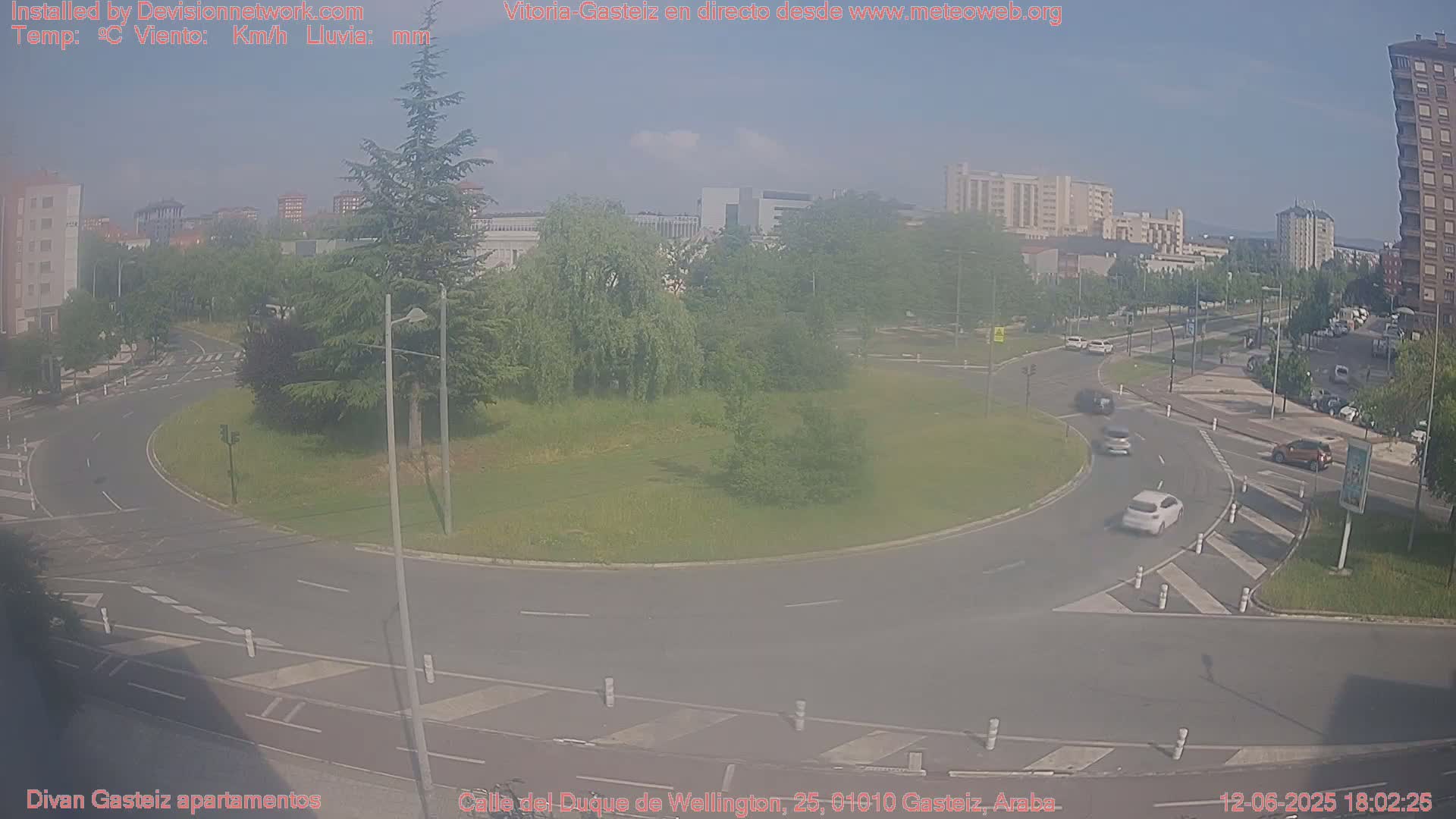 A hazy, sunny day shows a roundabout with several cars driving on it, surrounded by green grass, trees, and buildings in the background.