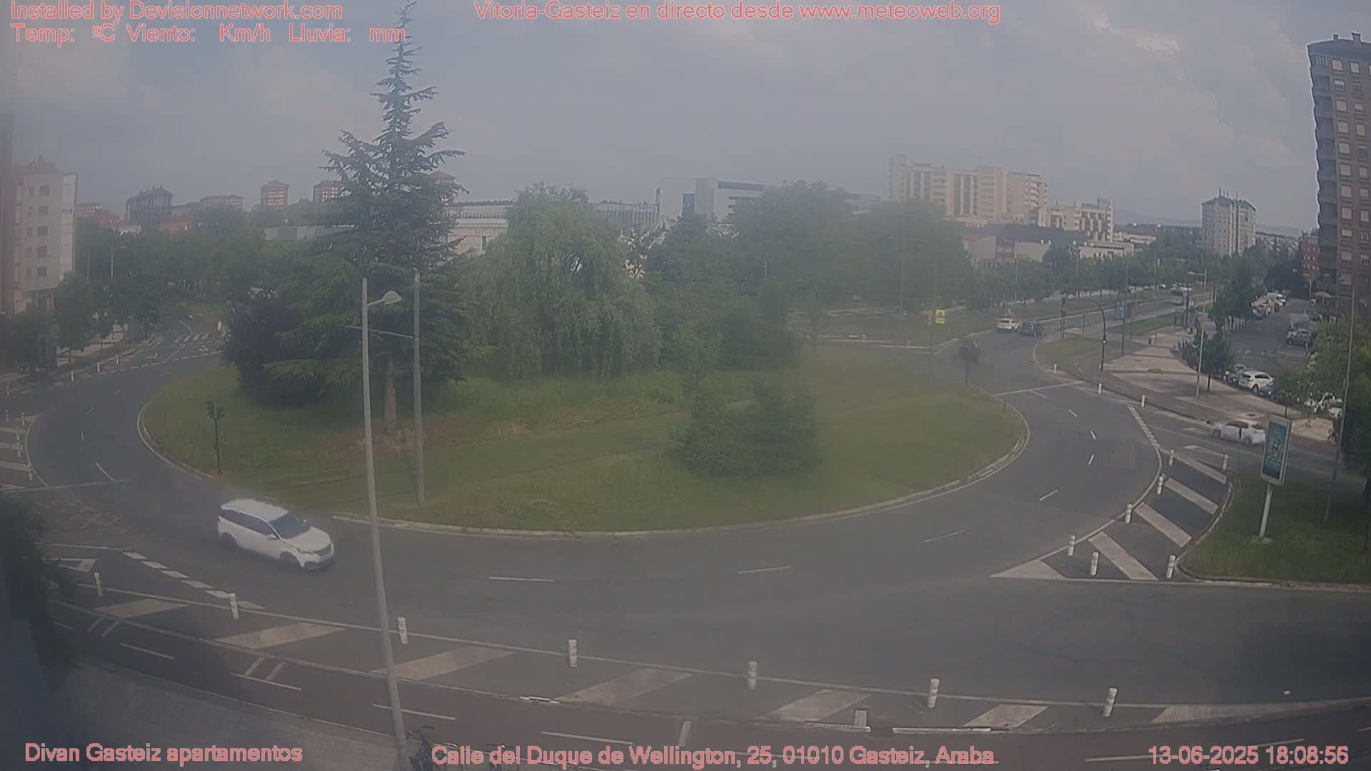 A hazy view of a roundabout with several cars and buildings in the background on a mostly cloudy day.