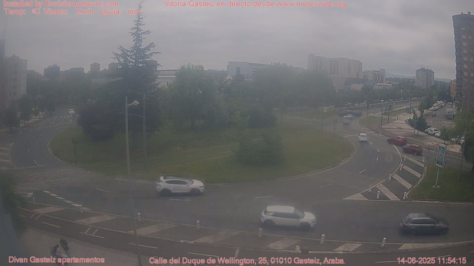 A hazy view of a roundabout with several cars, surrounded by green space and buildings in the background under overcast skies.