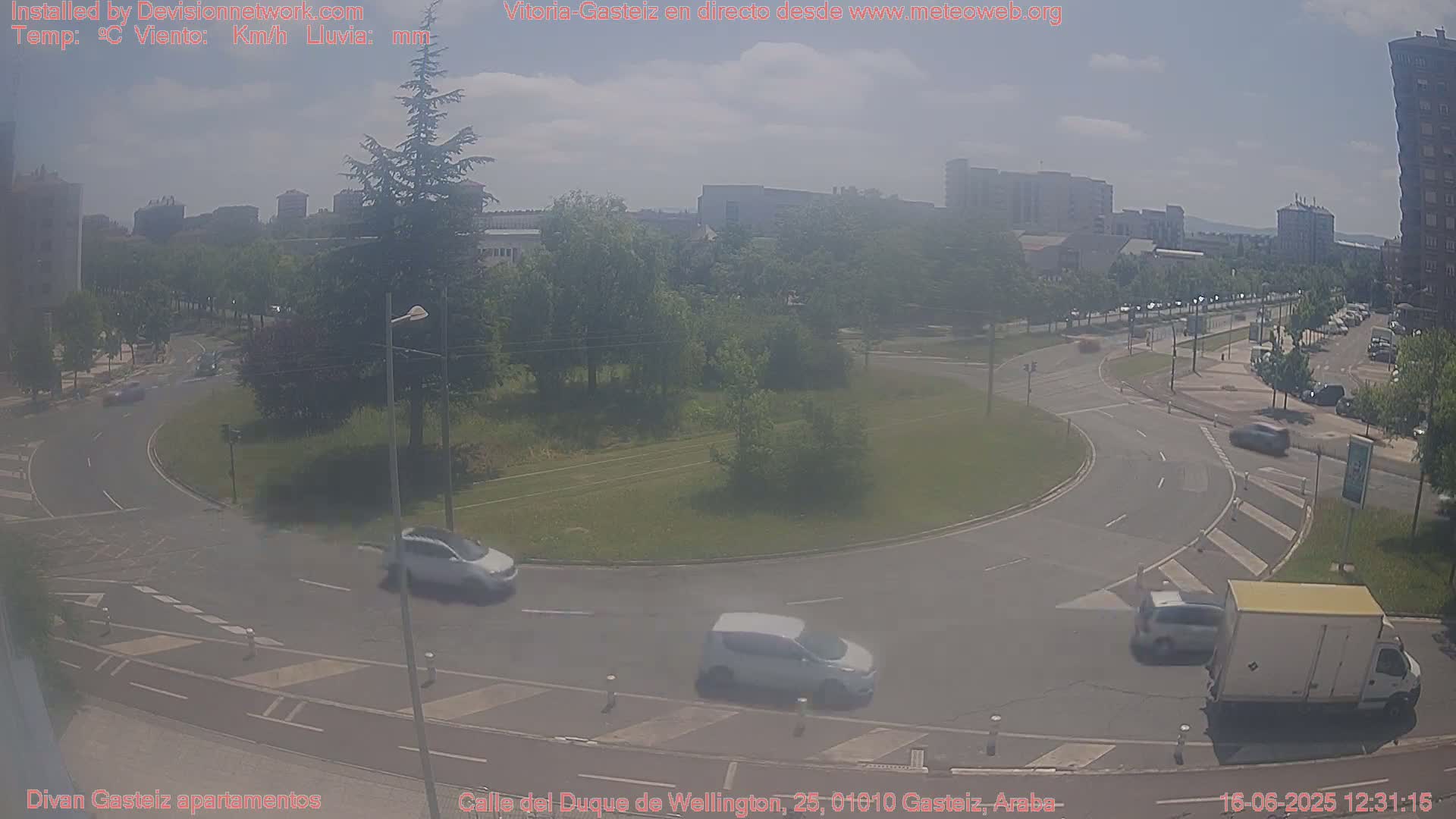 A hazy sunny day shows a roundabout in a city with several cars driving around it and buildings in the background.