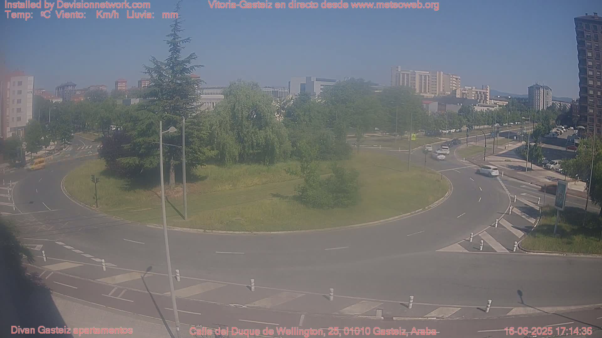 A sunny day shows a roundabout with several cars and buildings in the background, surrounded by green trees and grass.