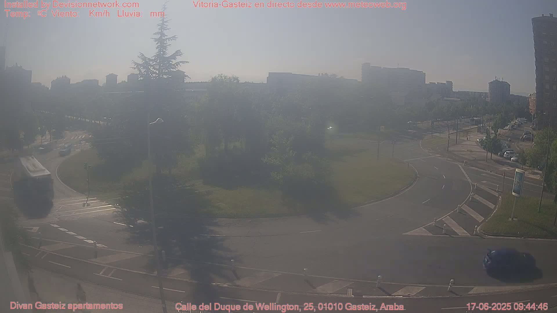 A hazy, sunny day shows a roundabout with several cars and a bus, surrounded by trees and buildings in the distance.