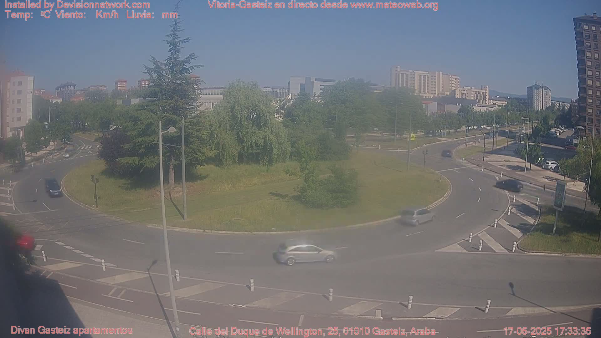 A sunny day shows a traffic roundabout in a city with several cars driving around it, and buildings in the background.