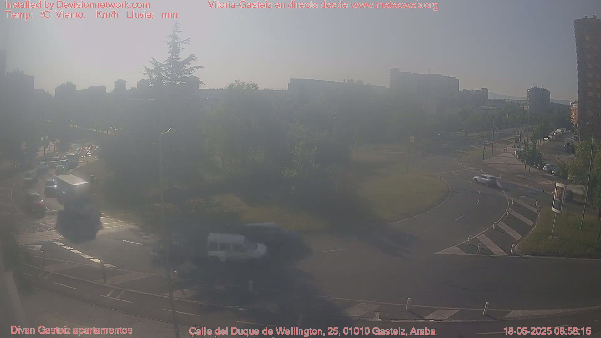 A hazy, sunny day shows a roundabout with several cars and buildings in the background.
