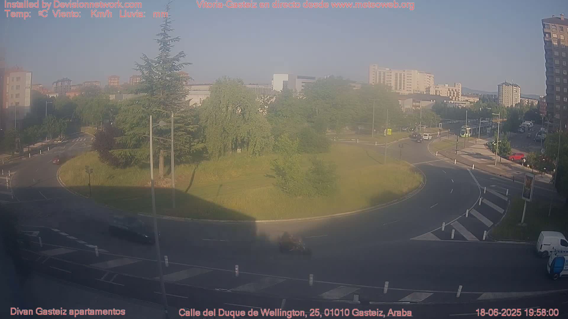 A hazy, sunny day shows a roundabout with several cars and a tram visible in the distance, surrounded by green trees and buildings.