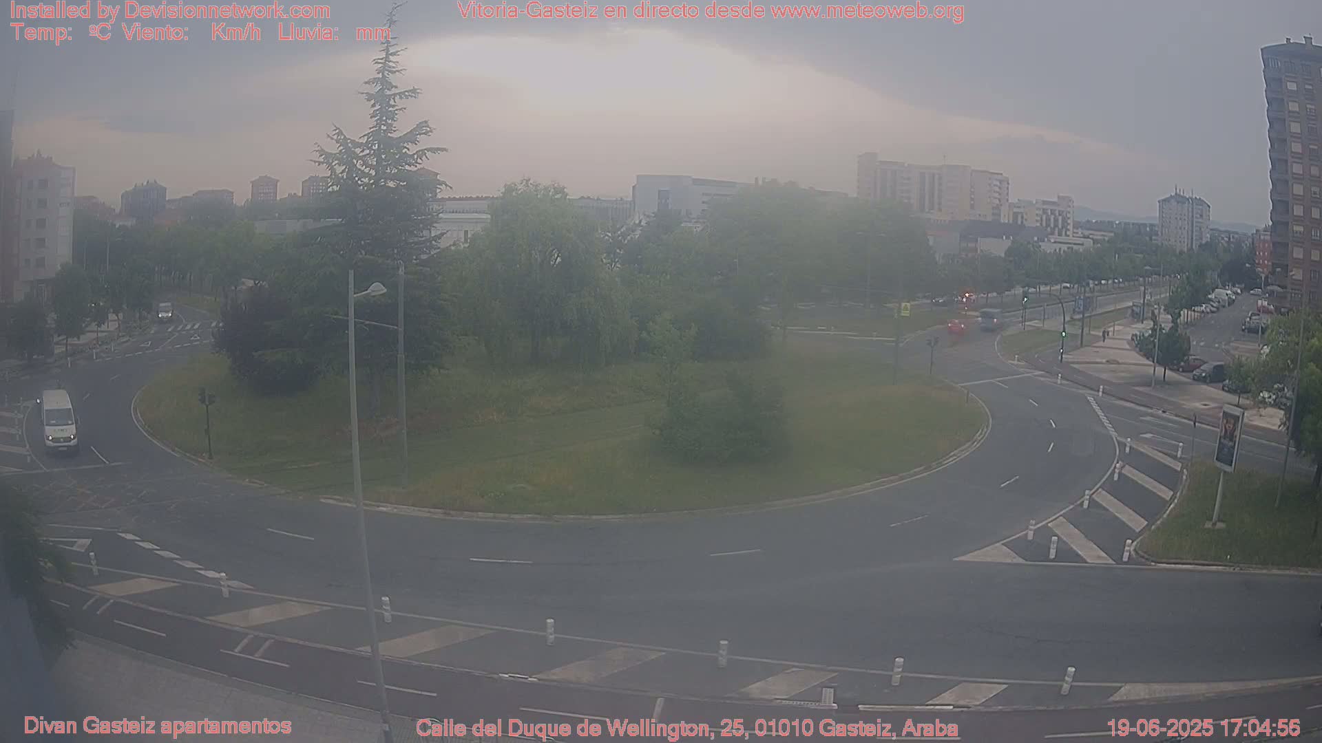 A hazy view of a roundabout in a city, with several cars and a van visible, under an overcast sky.