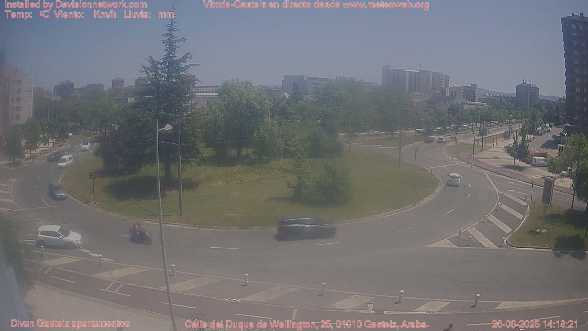 A hazy, sunny day shows a roundabout with several cars and a motorcycle navigating it, surrounded by buildings and green spaces.