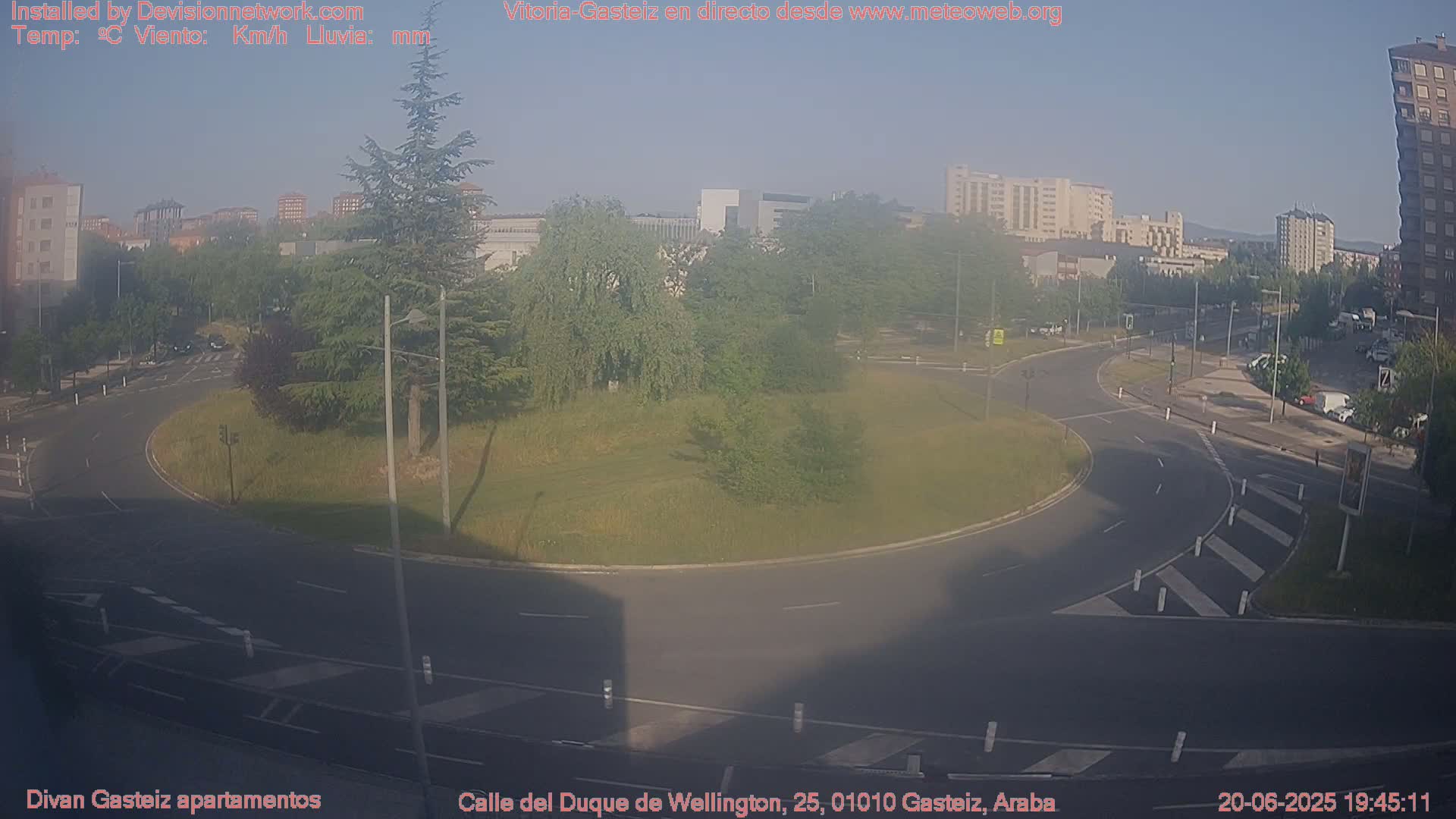 A mostly sunny view of a traffic circle surrounded by green grass and trees, with buildings and roads visible in the background.