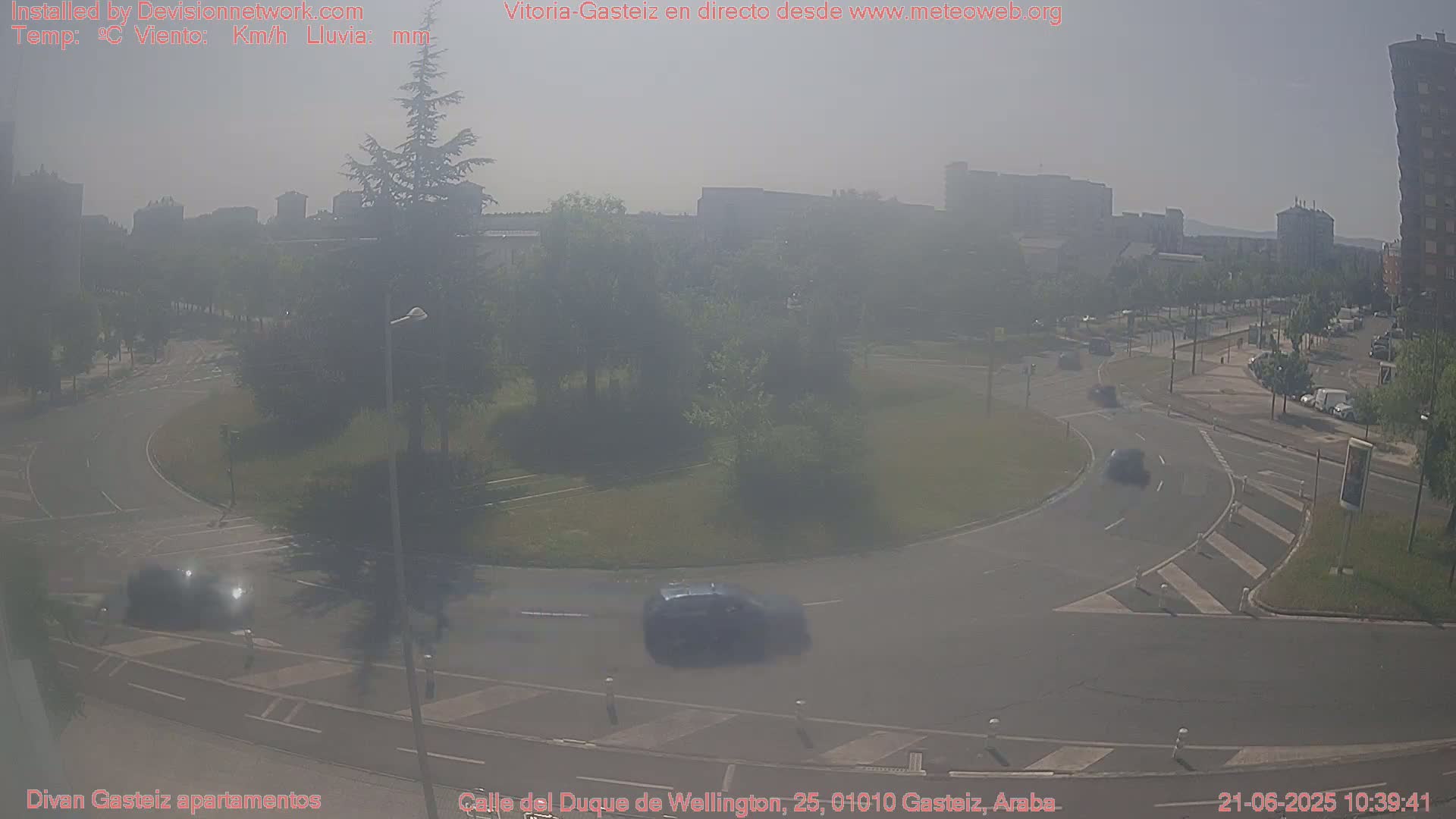 A hazy, sunny day reveals a roundabout with several cars driving through it and buildings in the background.