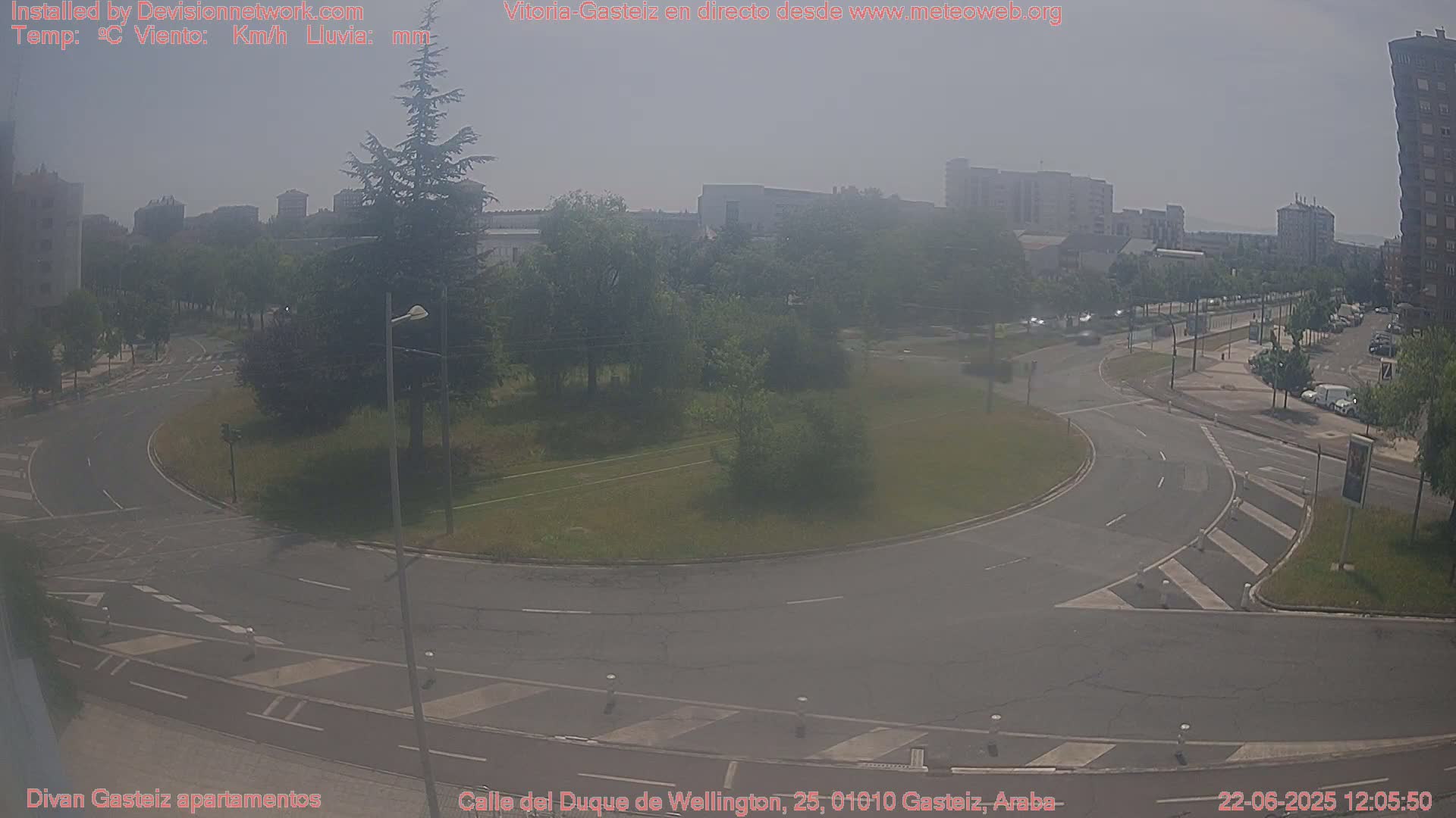 A hazy, sunny day reveals a roundabout and roadways surrounded by buildings and green spaces.