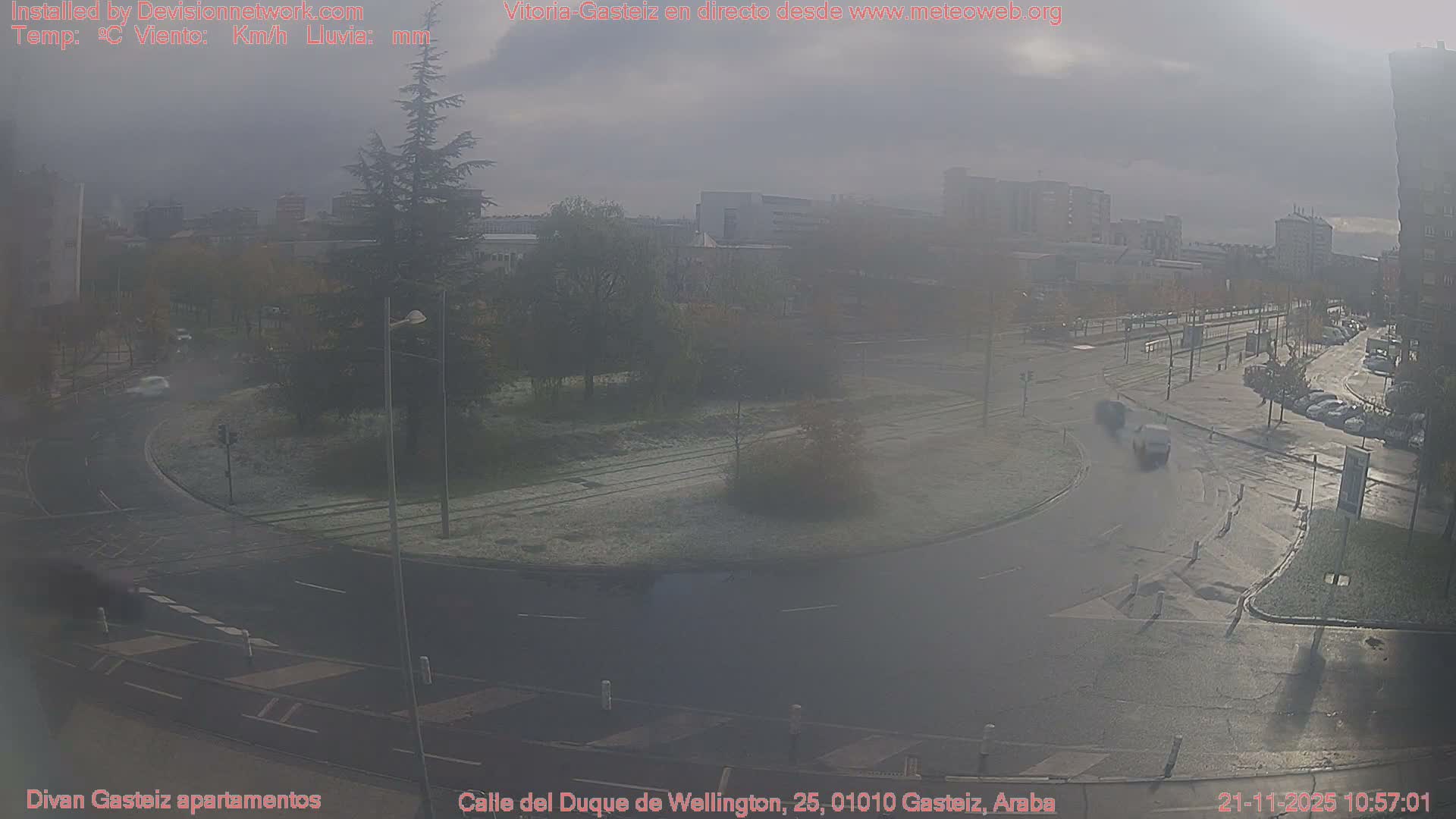 An overcast urban scene depicts a wide street intersection with cars, some in motion, and surrounding buildings amidst damp conditions and trees displaying hints of autumn.