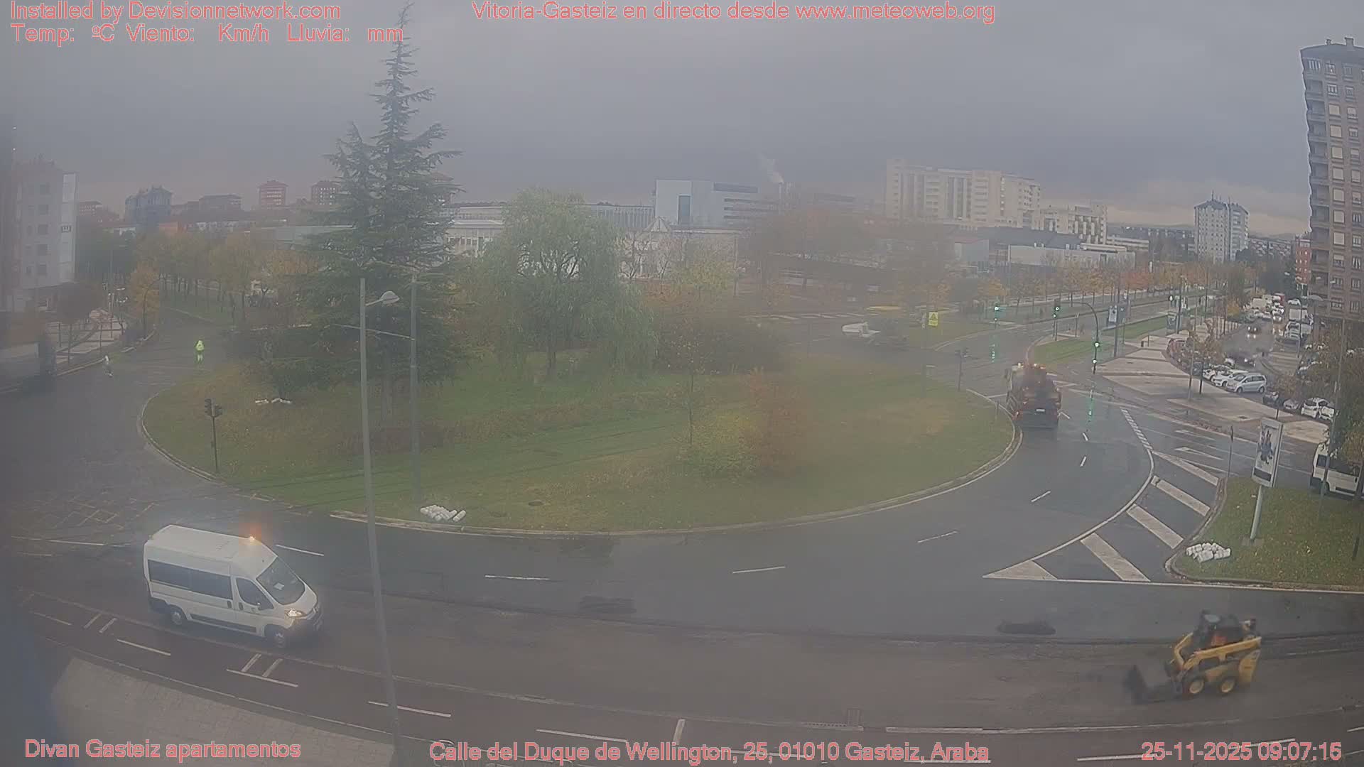 An overcast urban scene depicts wet roads with several vehicles, including a white van and a yellow construction vehicle, navigating a curve alongside grassy areas, trees, and distant buildings under a hazy grey sky.
