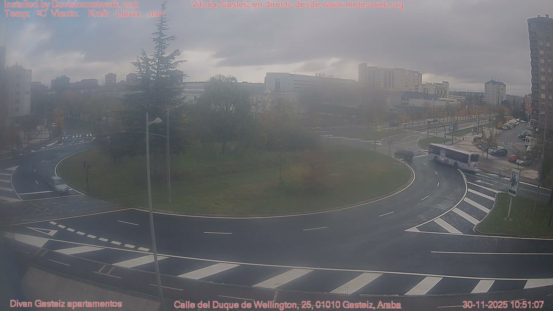 A grey, overcast day with wet roads shows a bus navigating a multi-lane roundabout in an urban area, with buildings and trees in the background.