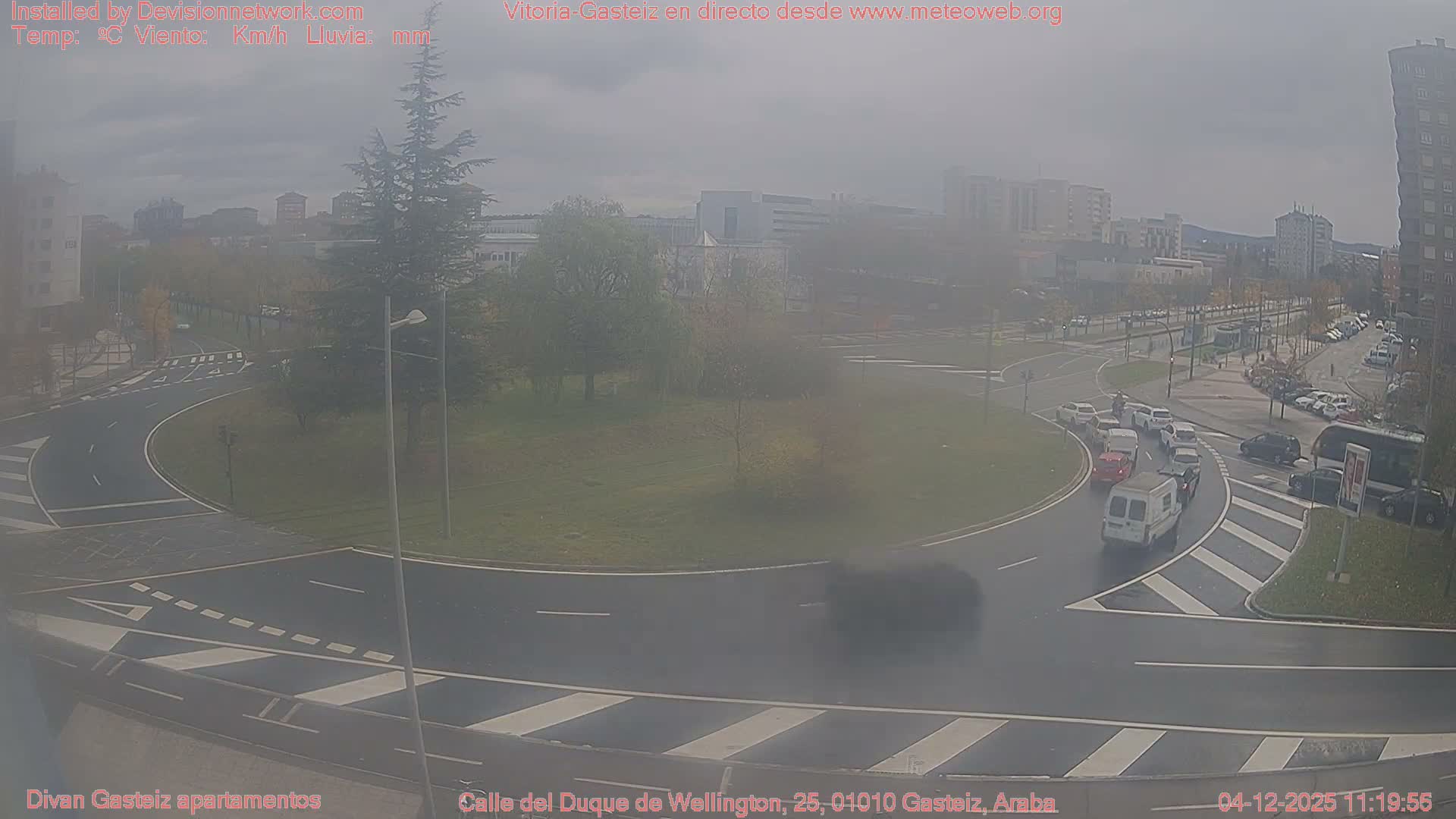 An elevated wide shot captures a multi-lane urban roundabout with wet asphalt, several cars in traffic, and surrounding buildings, all under a grey, overcast sky.
