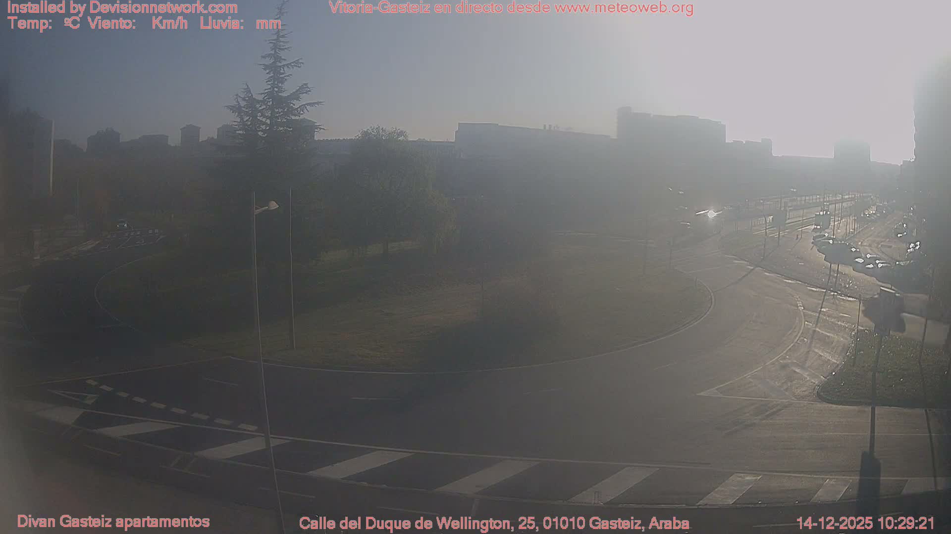 An elevated wide shot captures a multi-lane urban roundabout with wet asphalt, several cars in traffic, and surrounding buildings, all under a grey, overcast sky.