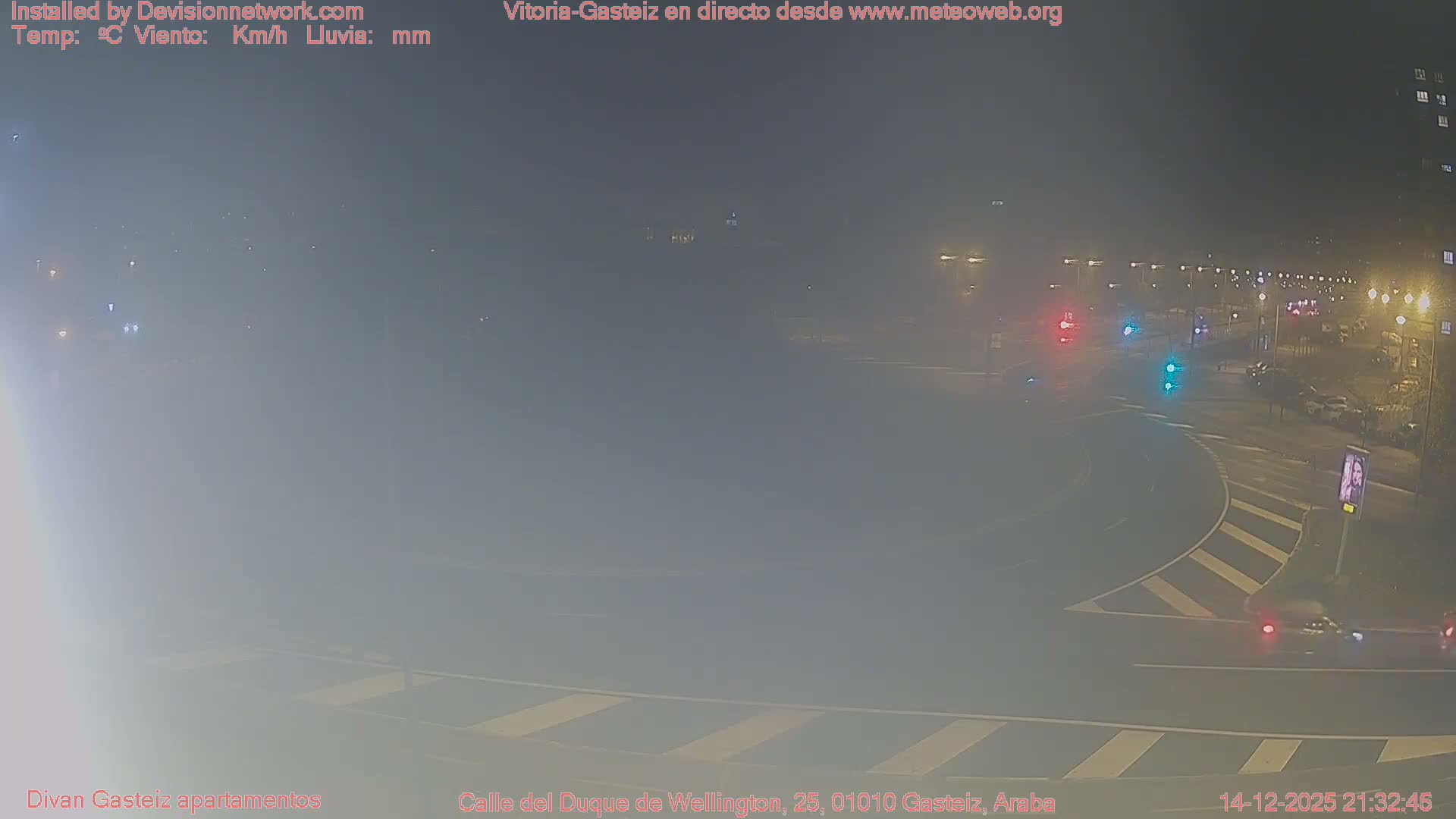 An elevated wide shot captures a multi-lane urban roundabout with wet asphalt, several cars in traffic, and surrounding buildings, all under a grey, overcast sky.