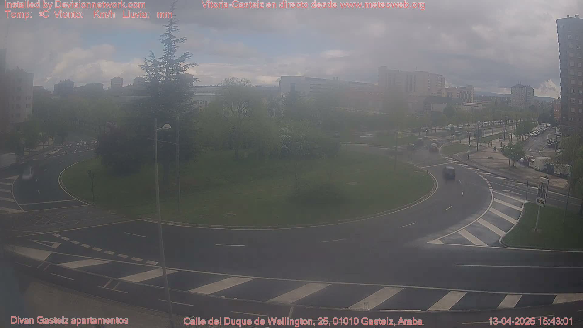 An elevated wide shot captures a multi-lane urban roundabout with wet asphalt, several cars in traffic, and surrounding buildings, all under a grey, overcast sky.