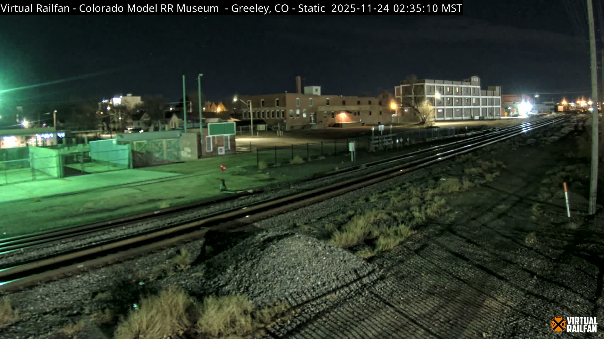 Multiple train tracks run through a well-lit industrial area at night, flanked by gravel, sparse vegetation, and various buildings under a clear, dark sky.
