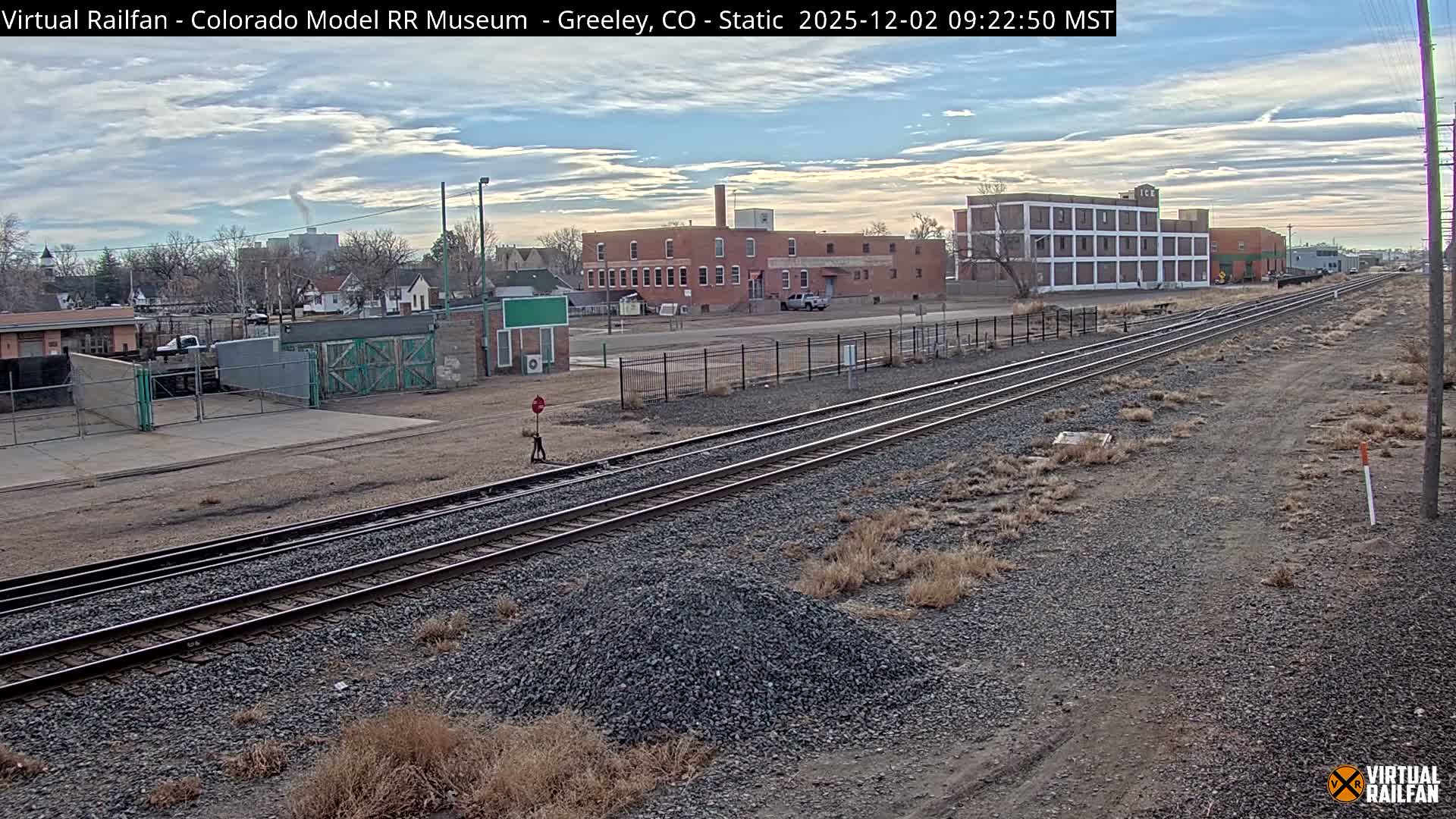 A wide view captures multiple railroad tracks stretching into the distance past various industrial buildings and a residential area, with sparse dry vegetation and gravel in the foreground, all under a partly cloudy sky.
