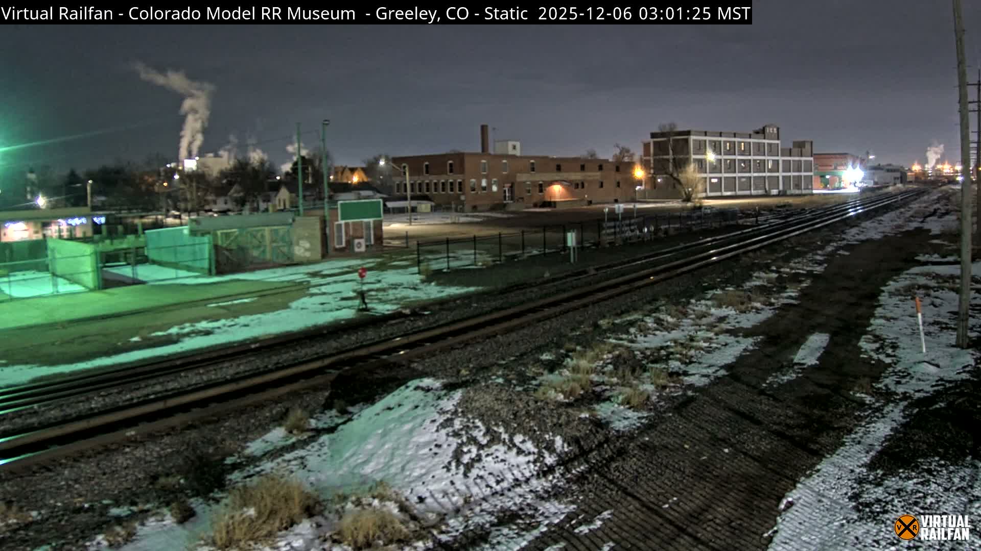 A nighttime view reveals a snowy railway yard with multiple tracks, flanked by illuminated industrial buildings and factories emitting plumes of steam into the cold, dark sky.