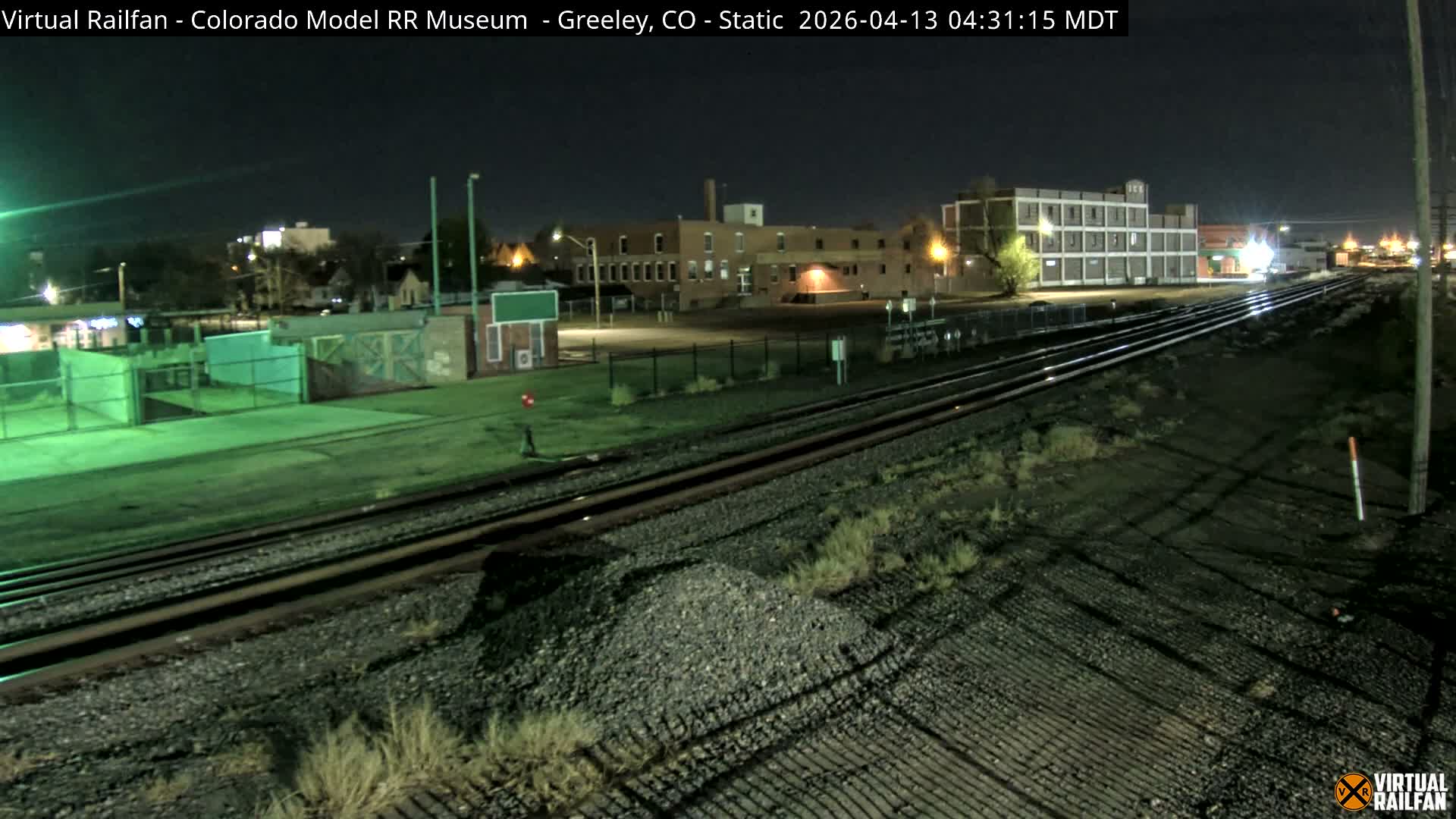 A nighttime view reveals a snowy railway yard with multiple tracks, flanked by illuminated industrial buildings and factories emitting plumes of steam into the cold, dark sky.