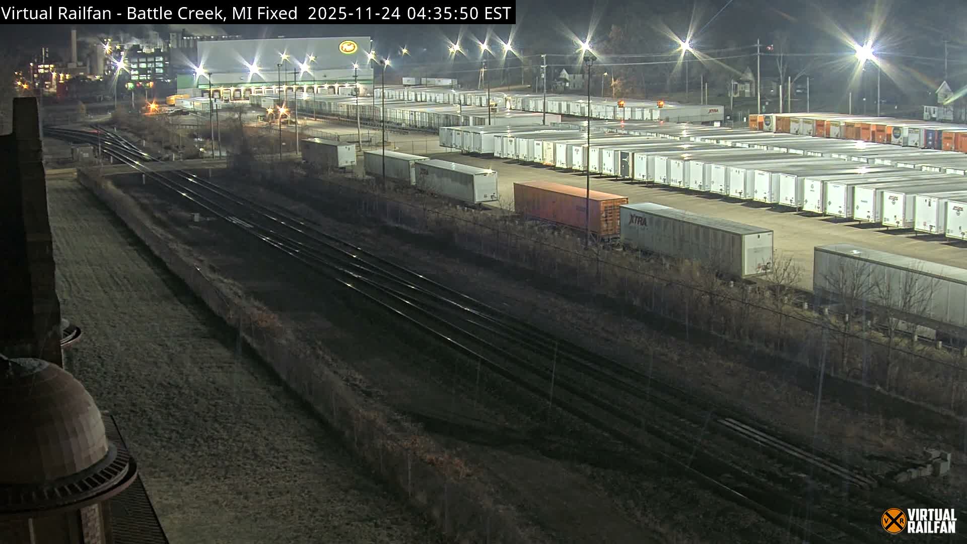 At night, an elevated view captures a large industrial scene featuring multiple train tracks alongside a sprawling yard filled with rows of semi-trailers and shipping containers, all brightly lit by numerous streetlights under clear skies.
