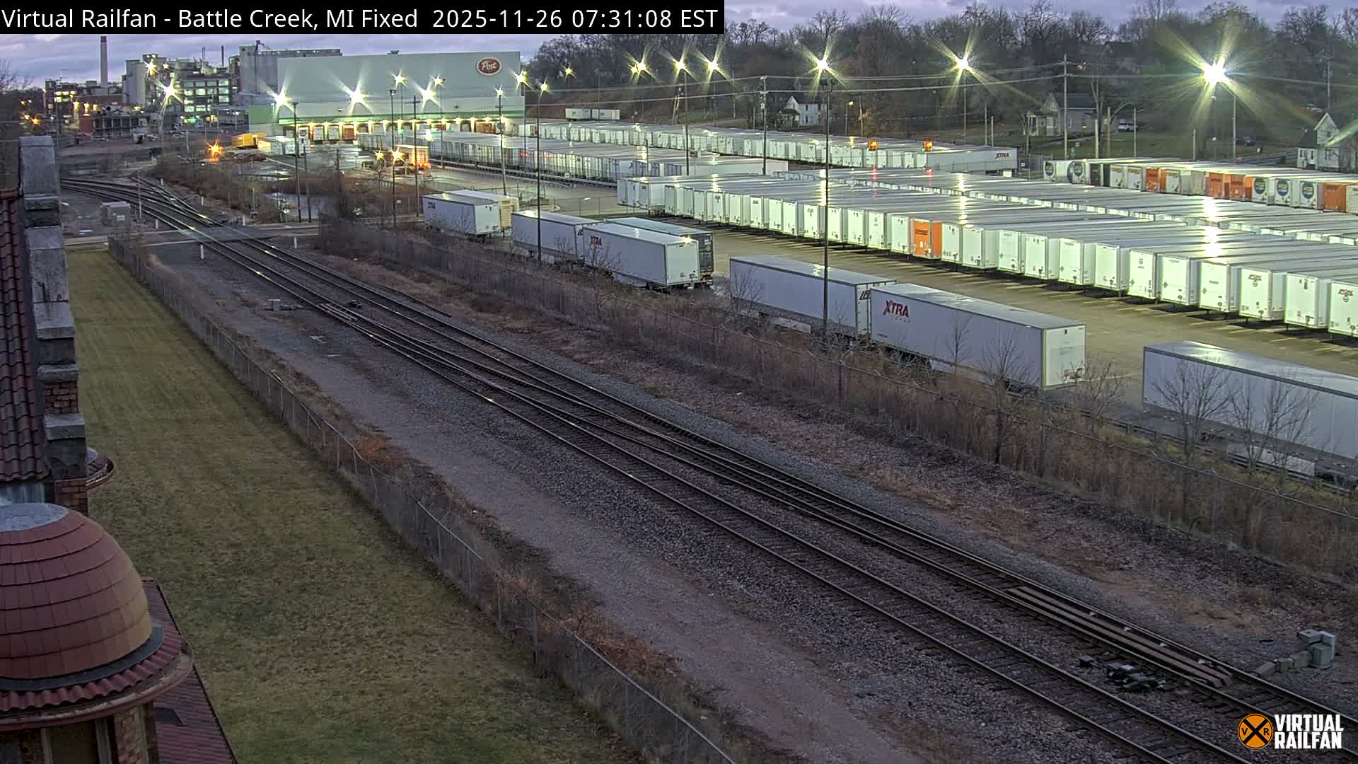 At night, an elevated view captures a large industrial scene featuring multiple train tracks alongside a sprawling yard filled with rows of semi-trailers and shipping containers, all brightly lit by numerous streetlights under clear skies.