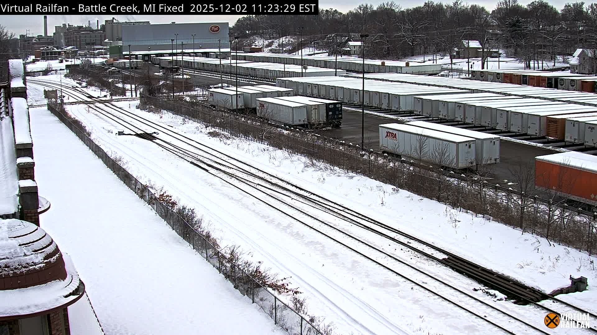 A snowy winter landscape features multiple train tracks running alongside a large yard filled with rows of snow-covered semi-trailers, with industrial buildings and bare trees visible under an overcast sky.