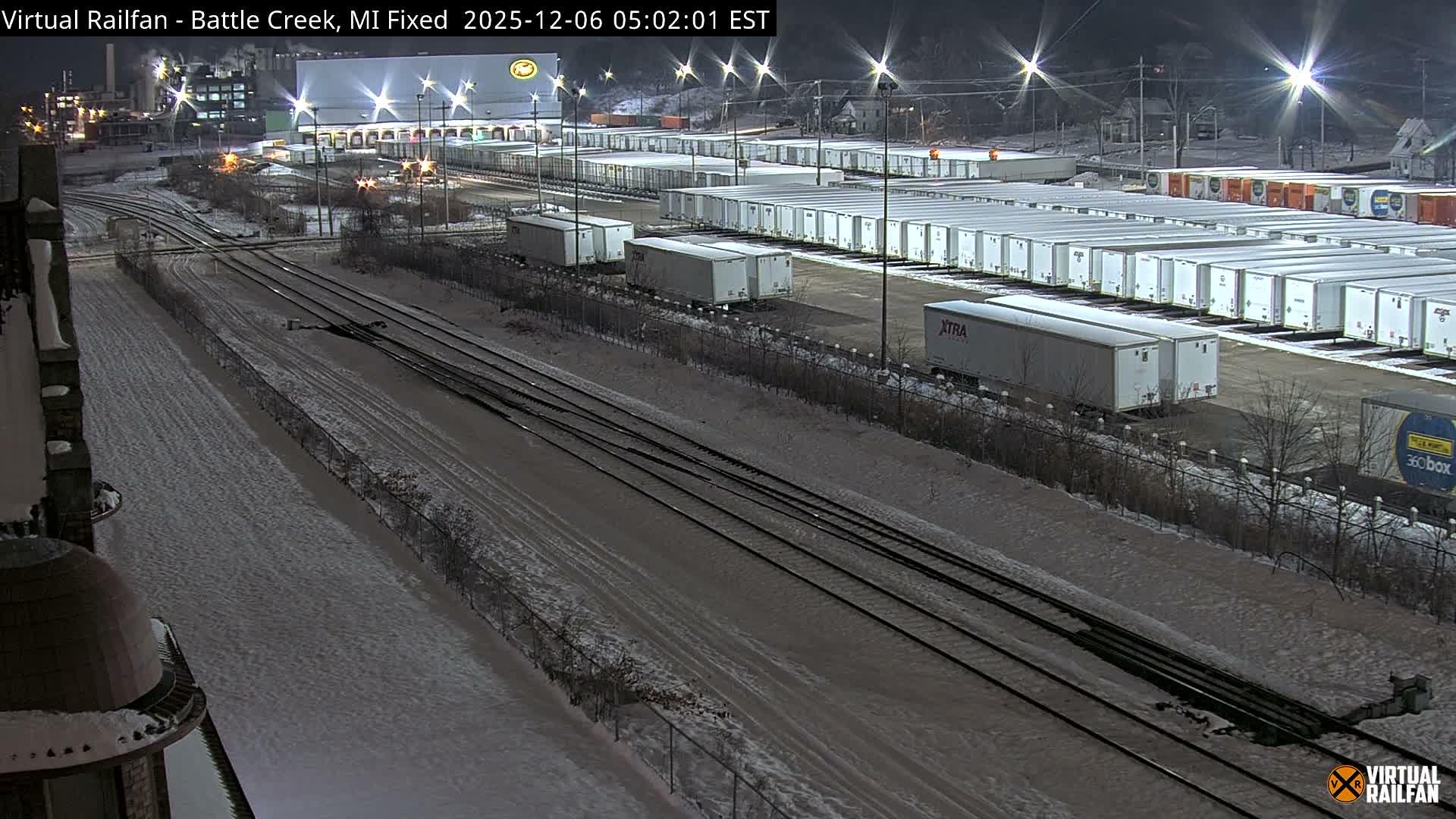 A nighttime view of a snow-covered industrial area features multiple railroad tracks in the foreground, a large lot filled with parked truck trailers, and brightly lit factory buildings under a dark sky.