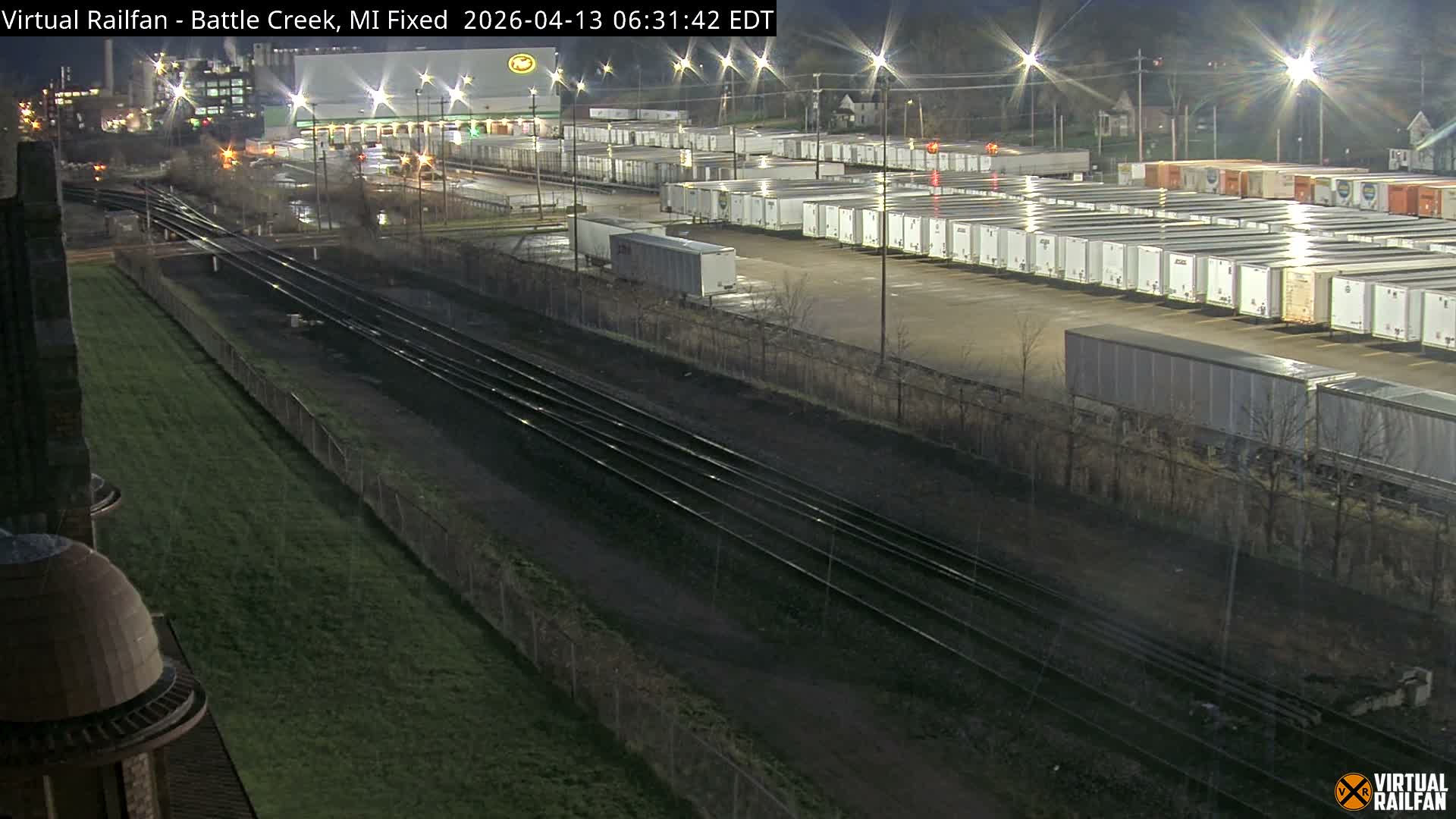 A nighttime view of a snow-covered industrial area features multiple railroad tracks in the foreground, a large lot filled with parked truck trailers, and brightly lit factory buildings under a dark sky.