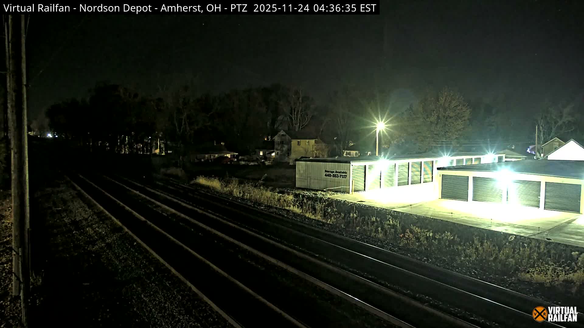A nighttime view features multiple railroad tracks receding into the distance, with brightly illuminated storage units and scattered houses on the right, and dark trees and a utility pole on the left, under clear conditions.