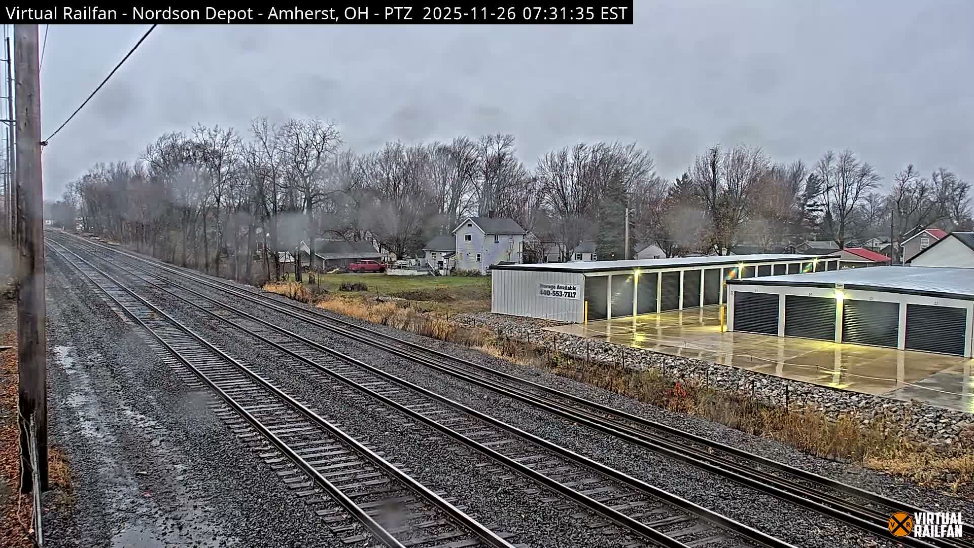 A nighttime view features multiple railroad tracks receding into the distance, with brightly illuminated storage units and scattered houses on the right, and dark trees and a utility pole on the left, under clear conditions.