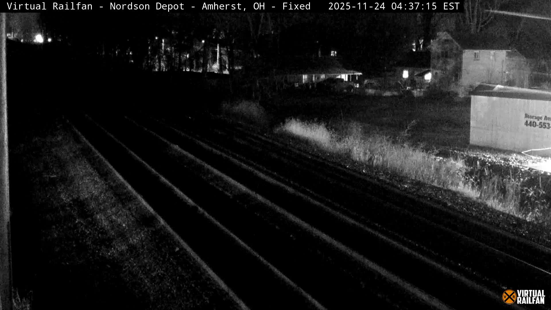 Multiple railroad tracks lead into the distance alongside an overgrown area, with dimly lit buildings and a storage container visible in the background under a clear night sky.